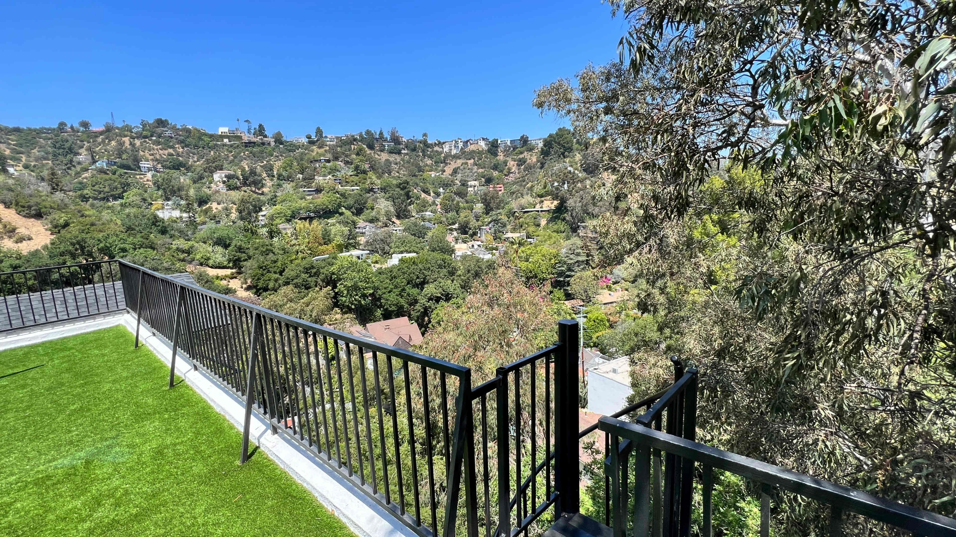 The image shows a balcony with a black railing overlooking a hillside covered in trees and houses under a clear blue sky.