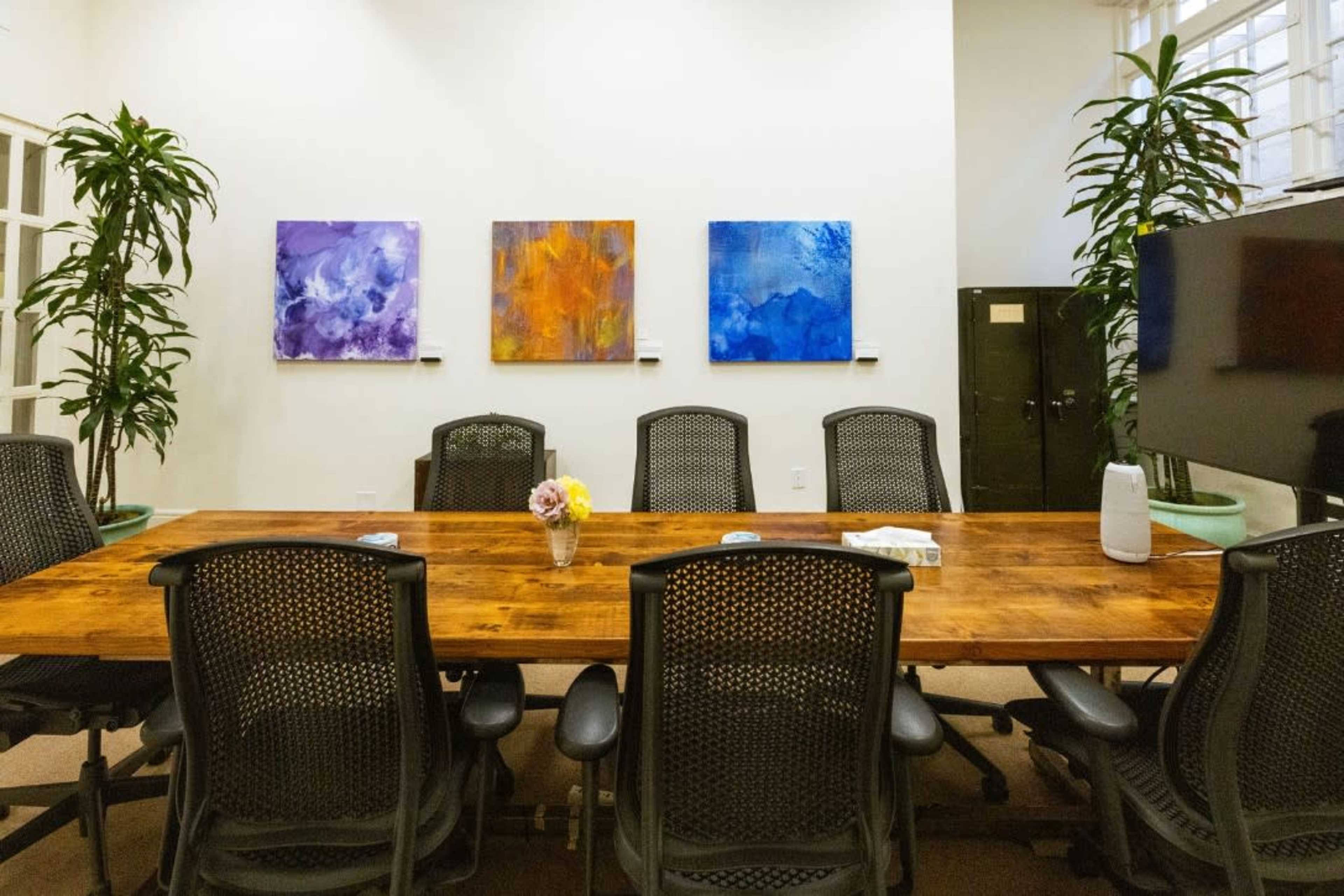 A conference room features a long wooden table surrounded by black office chairs, with colorful abstract paintings on the wall and tall plants in the corners.