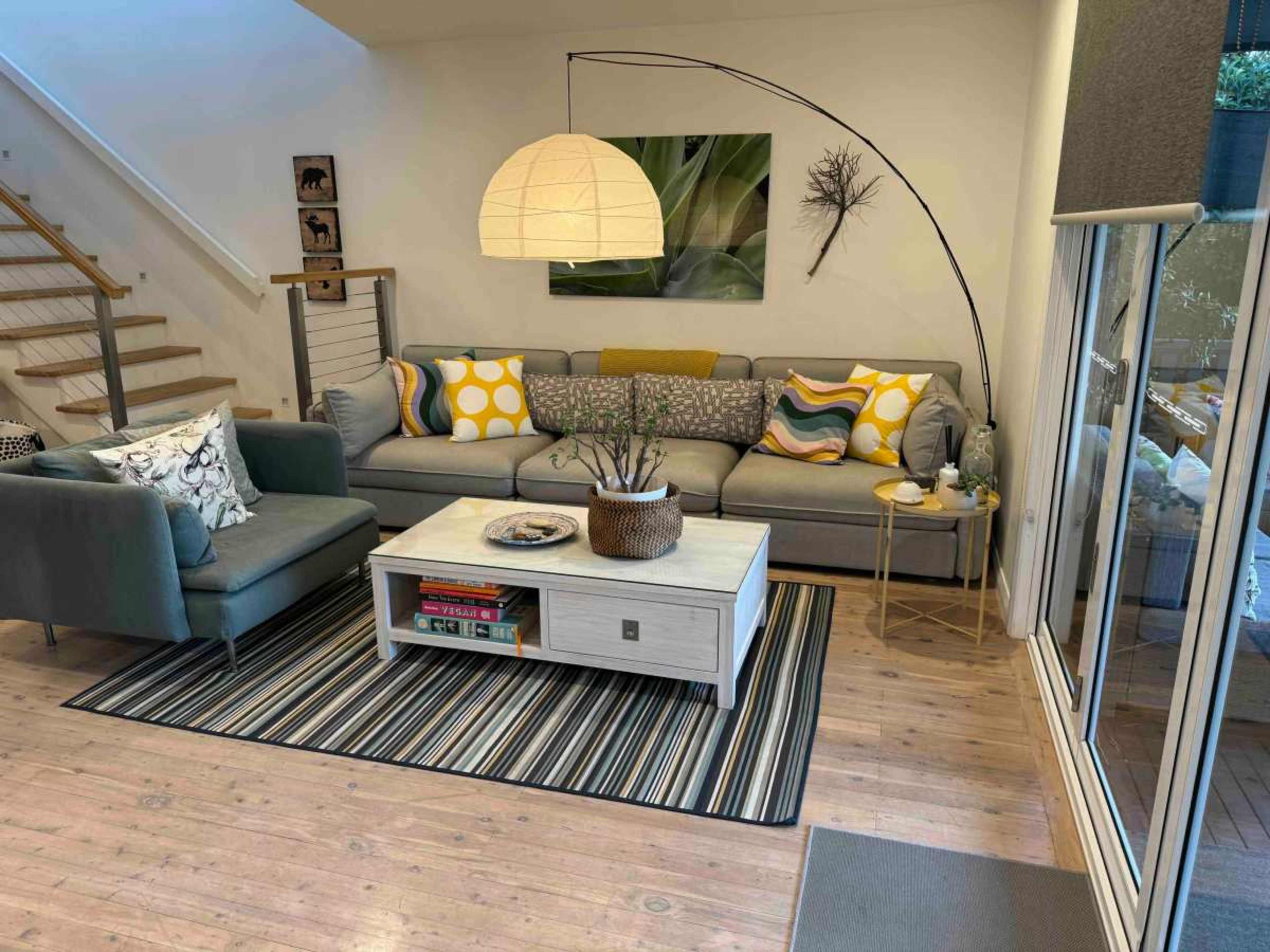 The image shows a modern living room with a gray sectional sofa, a coffee table, and a large pendant light, complemented by a striped rug and a staircase in the background.