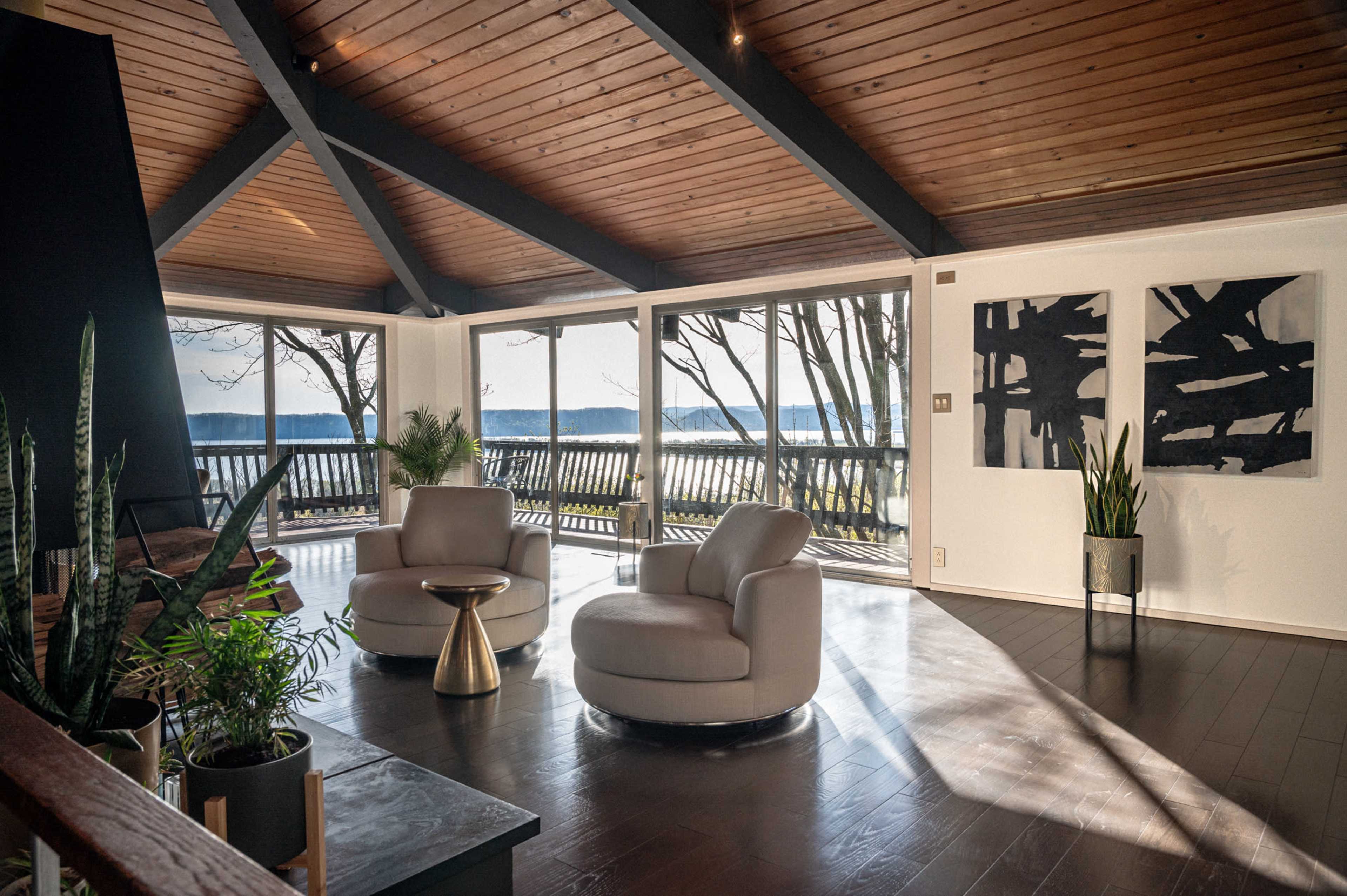 A modern living room with two white armchairs, a small side table, large windows overlooking a scenic view, and decorative plants.
