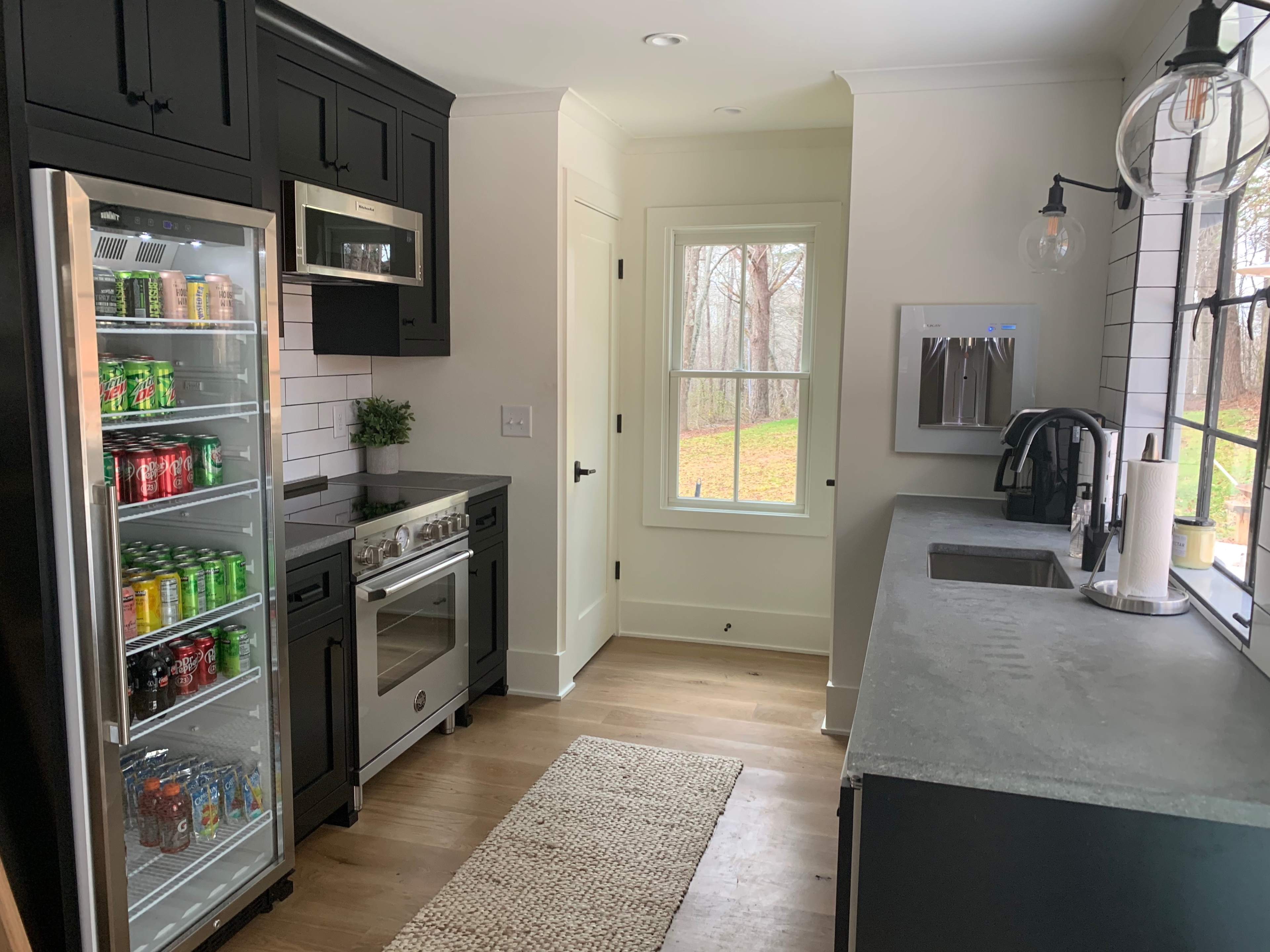 A modern kitchen features black cabinetry, a glass-front refrigerator filled with beverages, and stainless steel appliances alongside a window with a view of a wooded area.