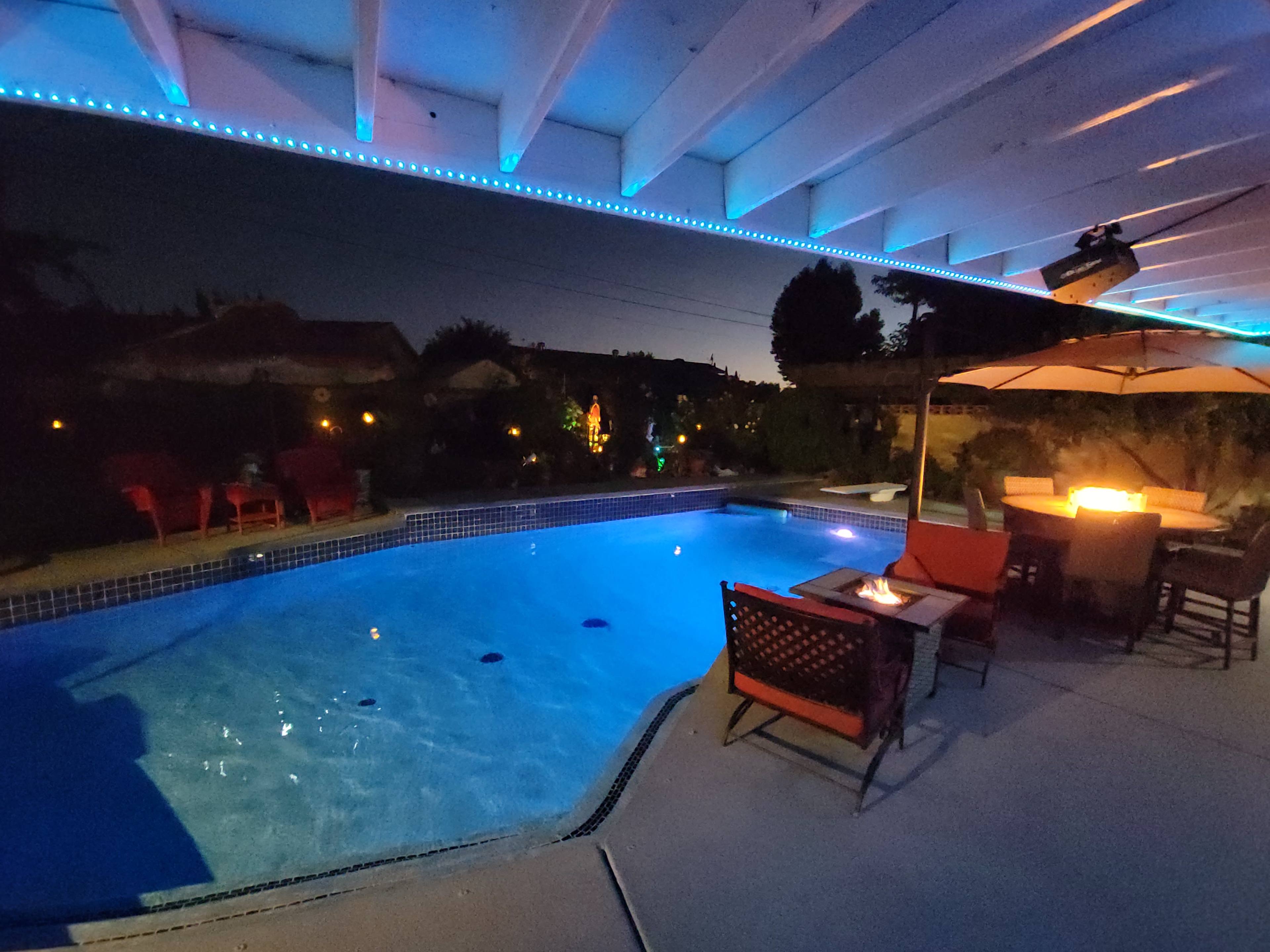 A well-lit pool area features a vibrant blue pool under a patio with outdoor seating and an umbrella.