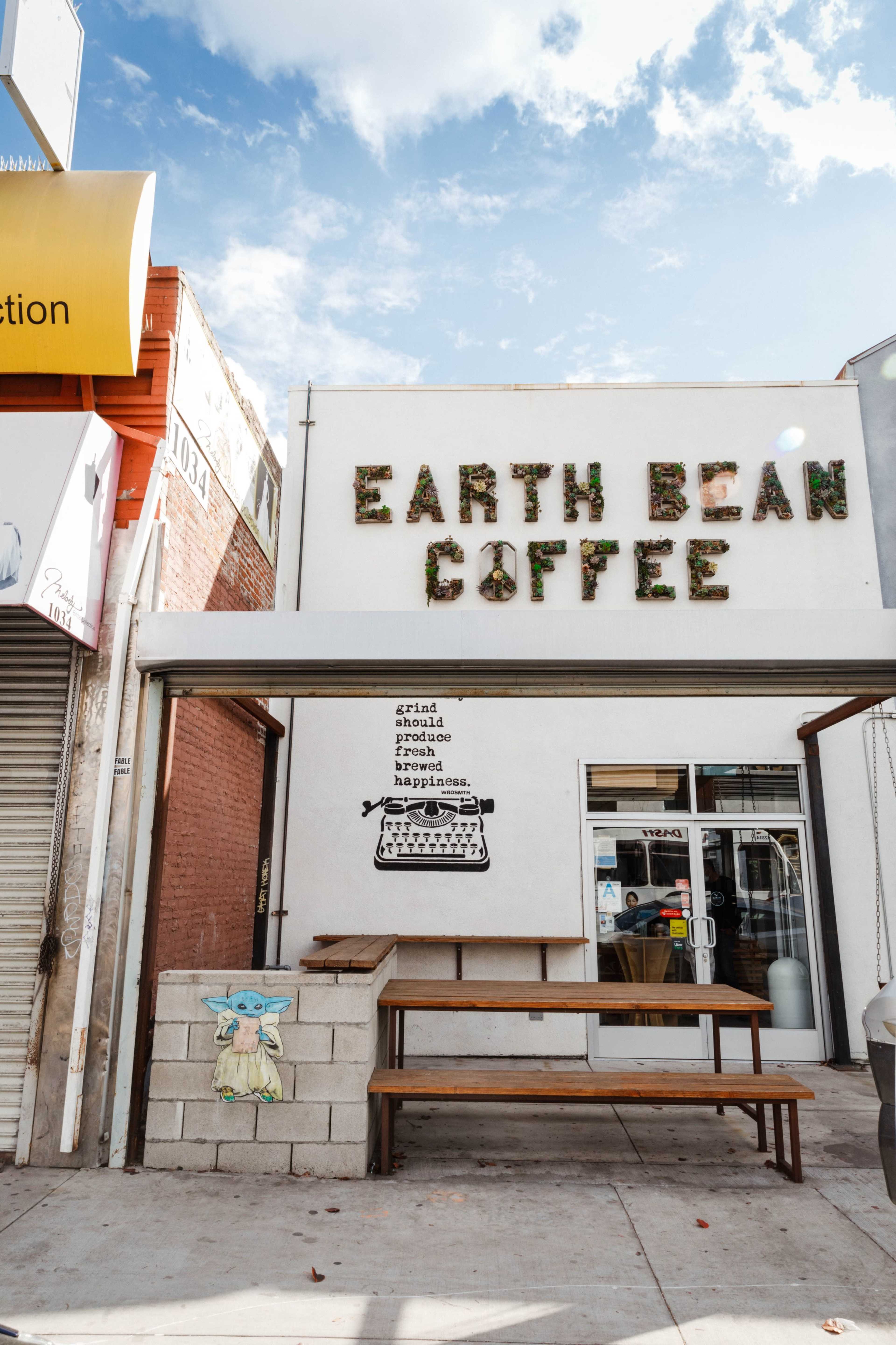 The image shows the exterior of a coffee shop named "Earth Bean Coffee," featuring a wooden table in front and a mural of a typewriter on the wall.