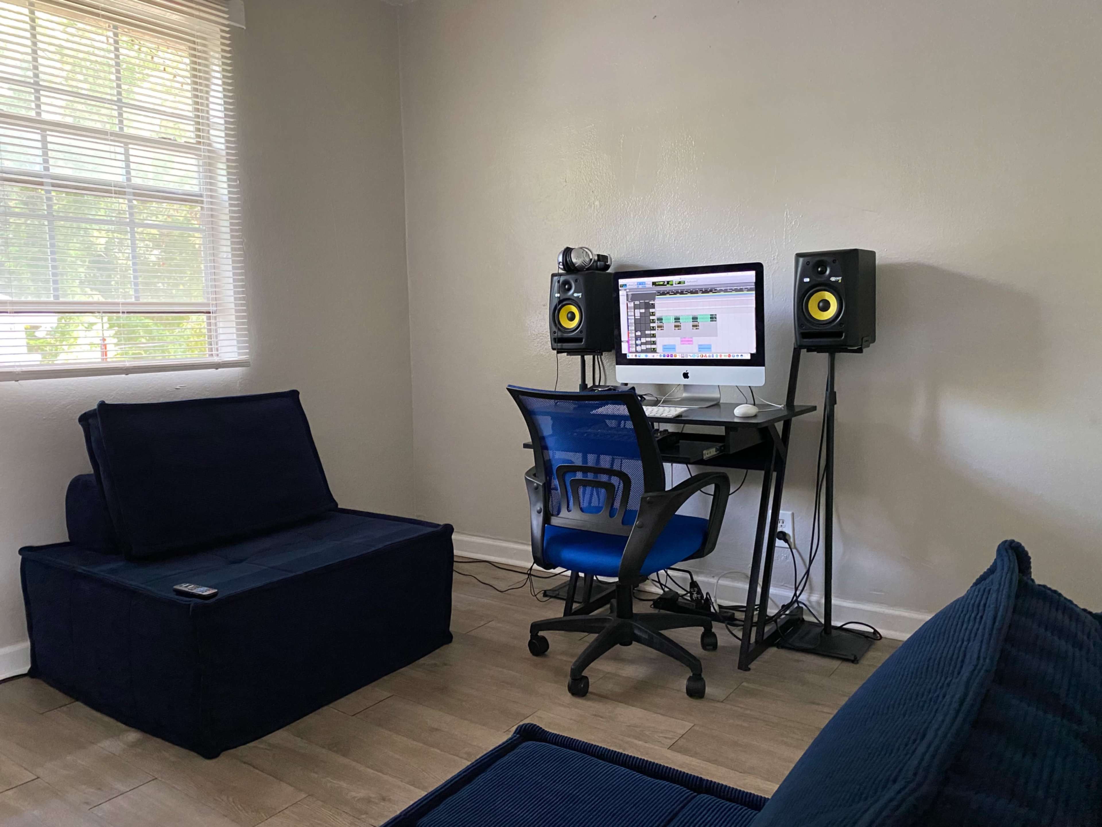 The image shows a small home office setup with an iMac on a desk, flanked by studio monitors, and a blue seating area.