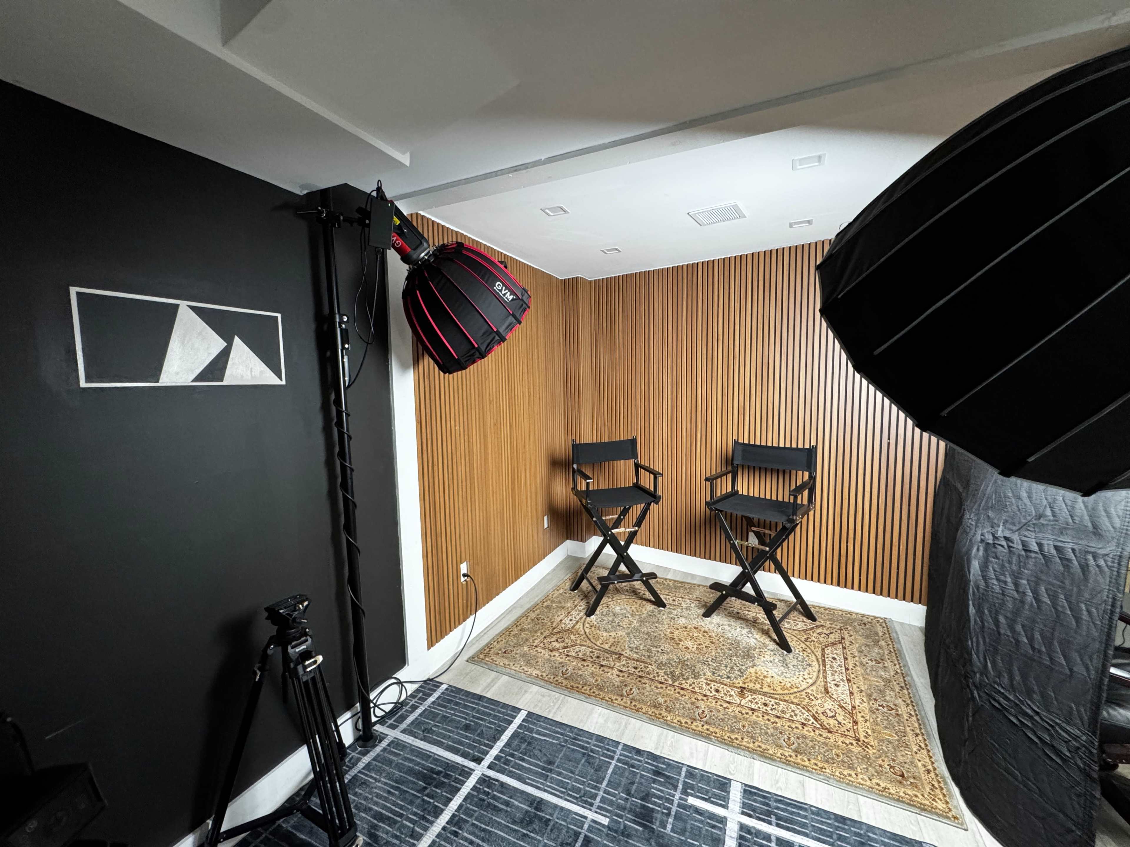 The image shows a photography studio setup with two director's chairs, a patterned rug, and professional lighting equipment against a wooden accent wall.
