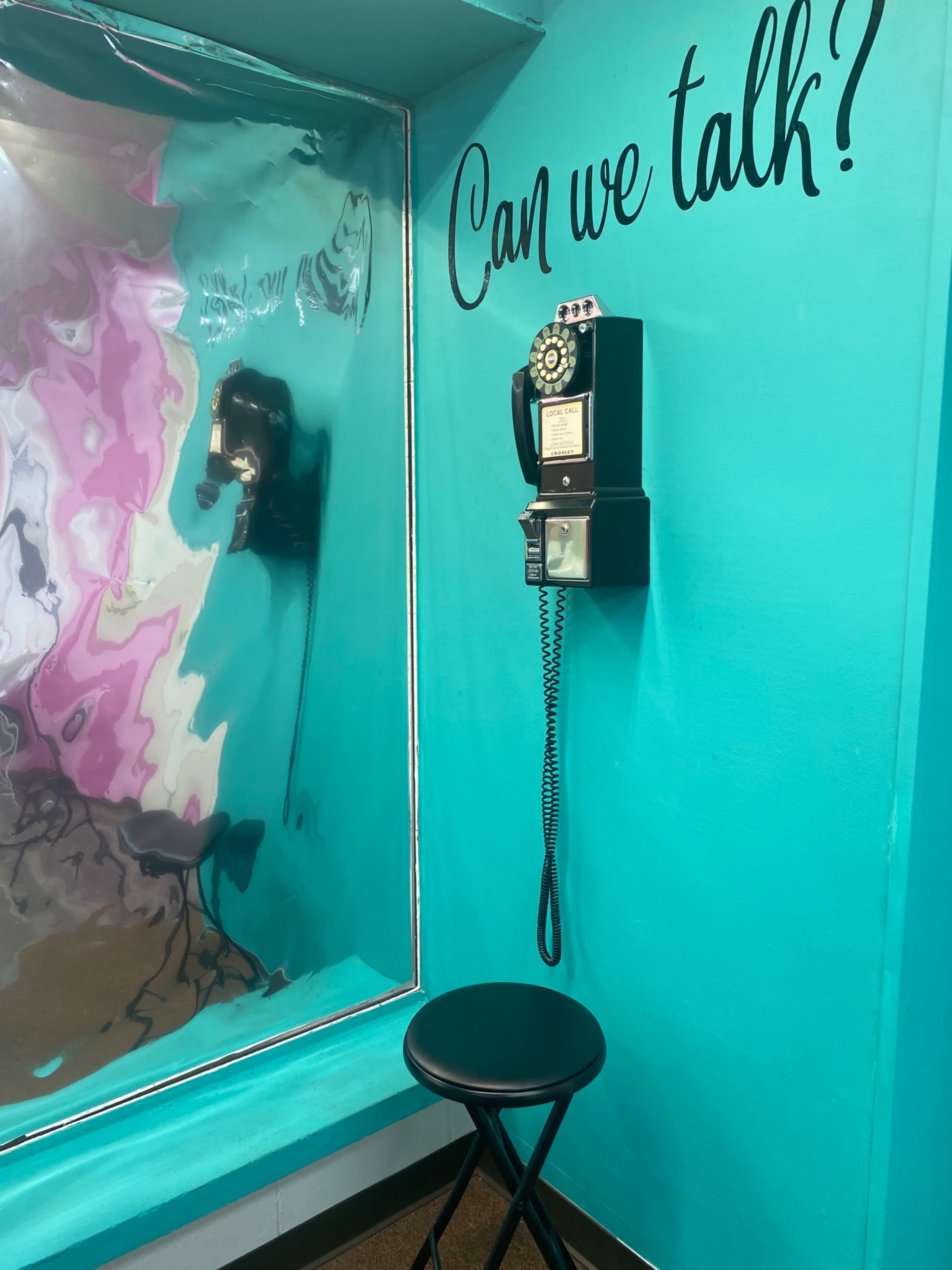 A vintage payphone is mounted on a bright turquoise wall next to a reflective metallic surface, accompanied by the text "Can we talk?" above it.