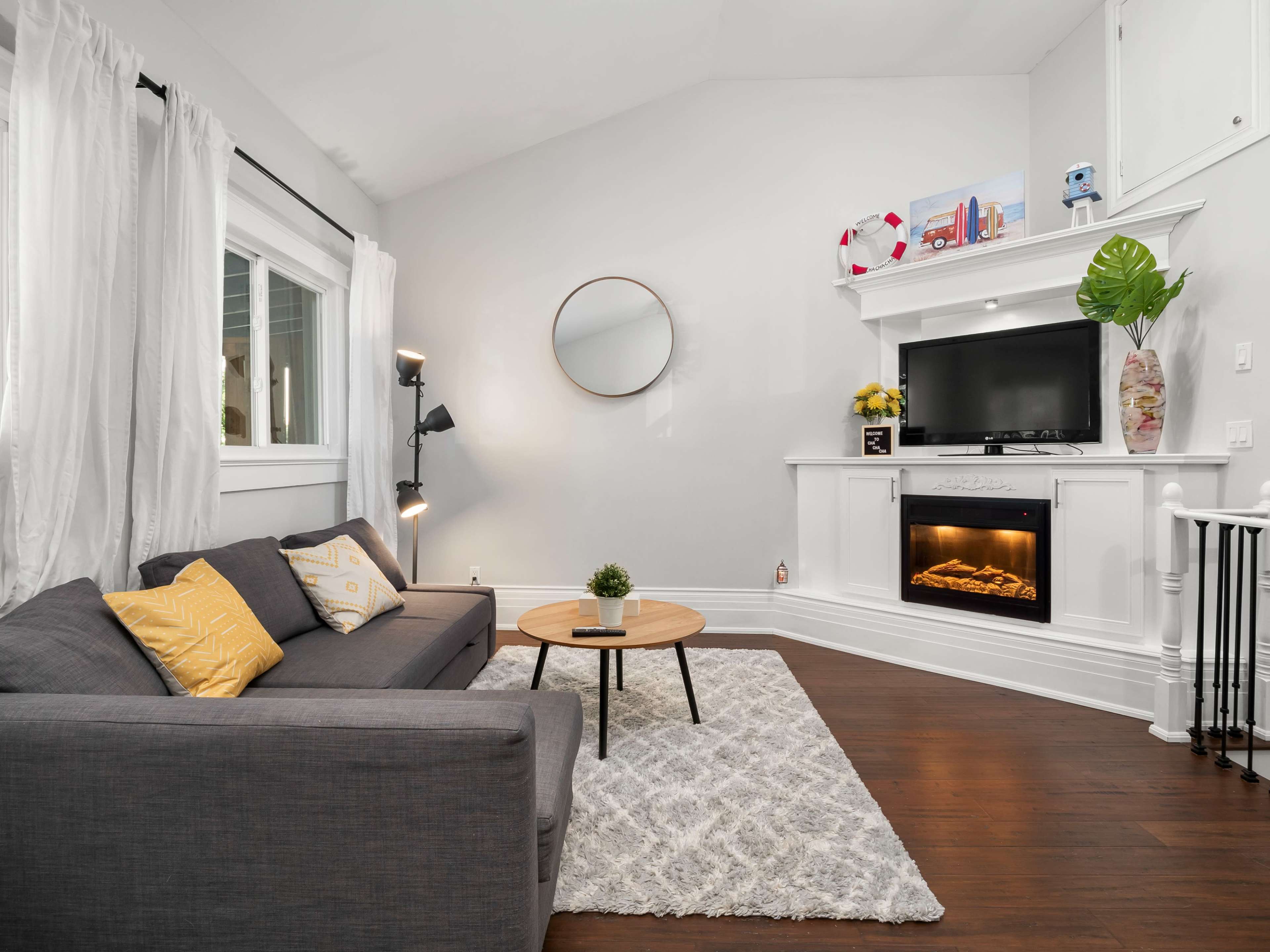 A modern living room features a gray sofa, a round coffee table, a wall-mounted television, and an electric fireplace, all decorated with light-colored curtains and a circular mirror.