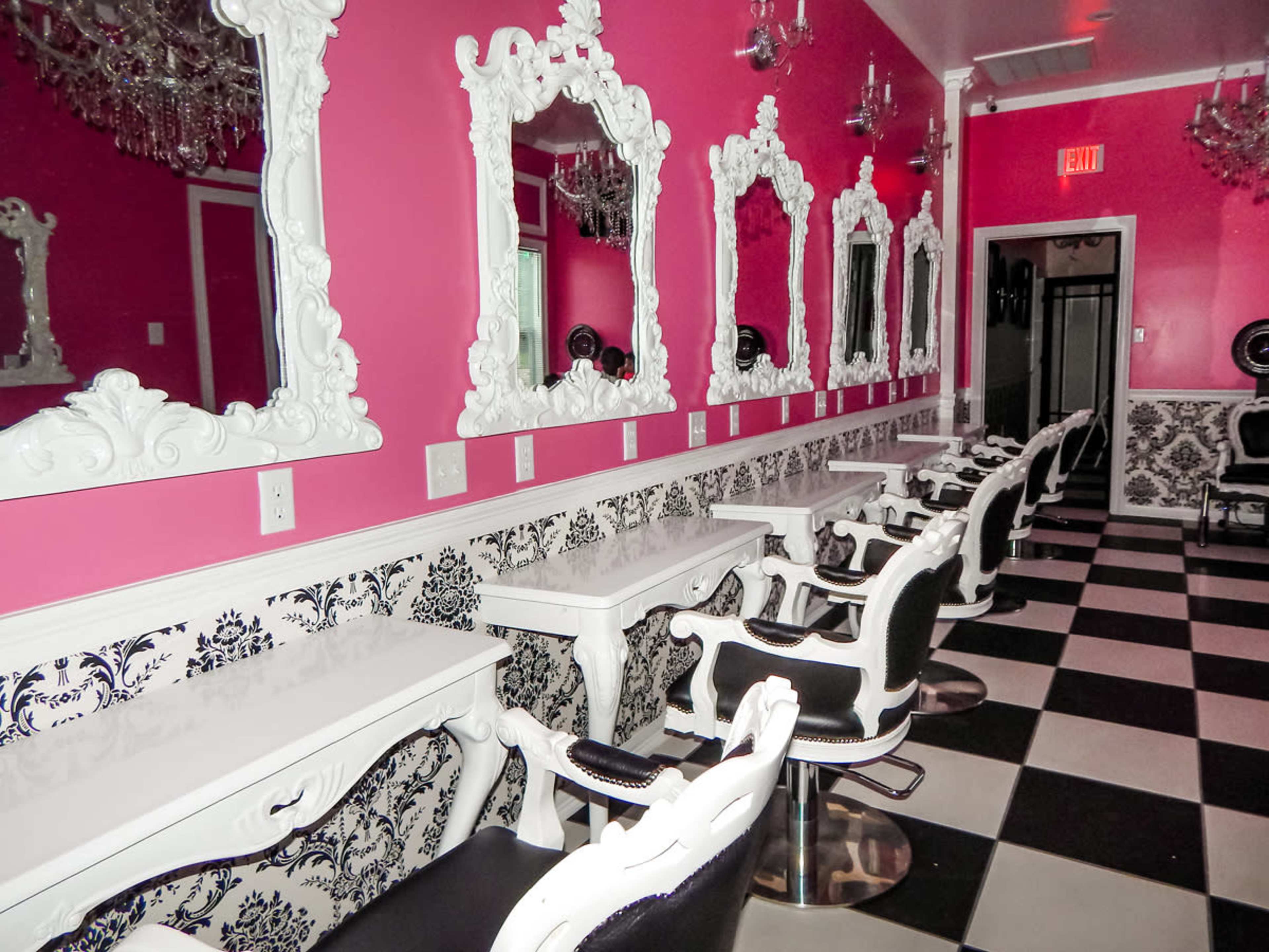 A brightly colored salon with white and black striped flooring, ornate white mirrors, and black salon chairs arranged along a pink wall.