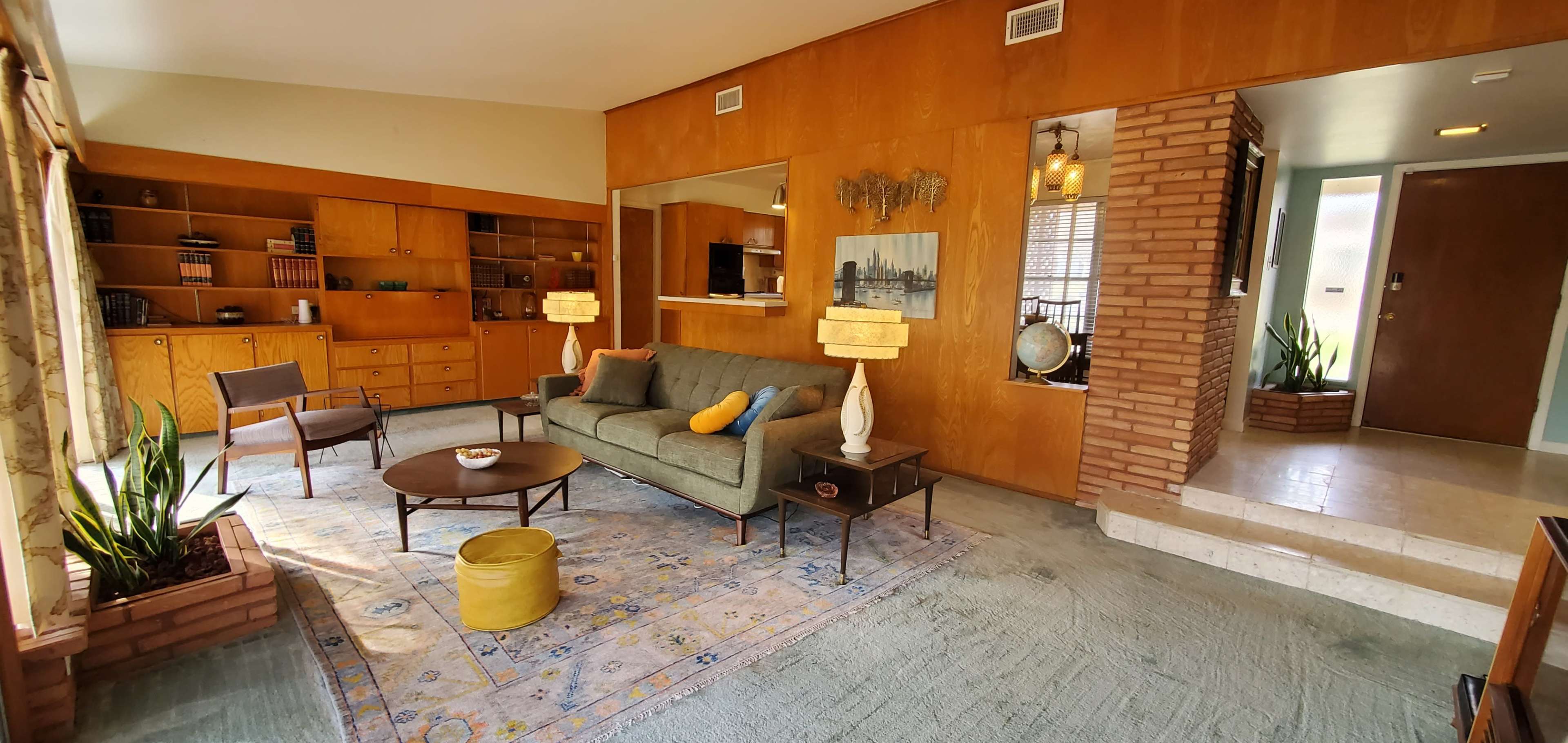 A mid-century modern living room featuring a green sofa, wooden paneling, a round coffee table, and decorative lamps, with a small staircase leading to a door.