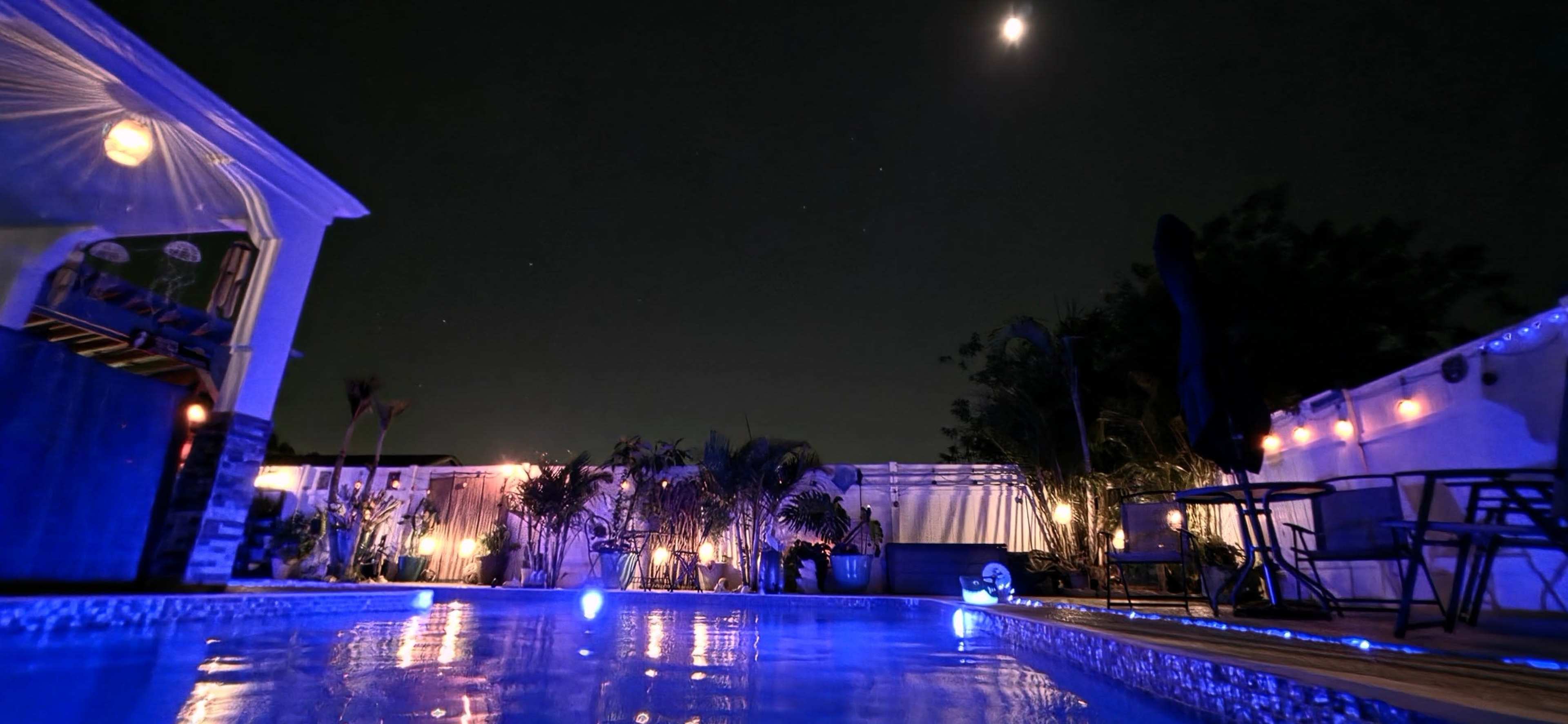 The image shows a softly illuminated swimming pool surrounded by plants and outdoor furniture under a night sky with a visible moon.