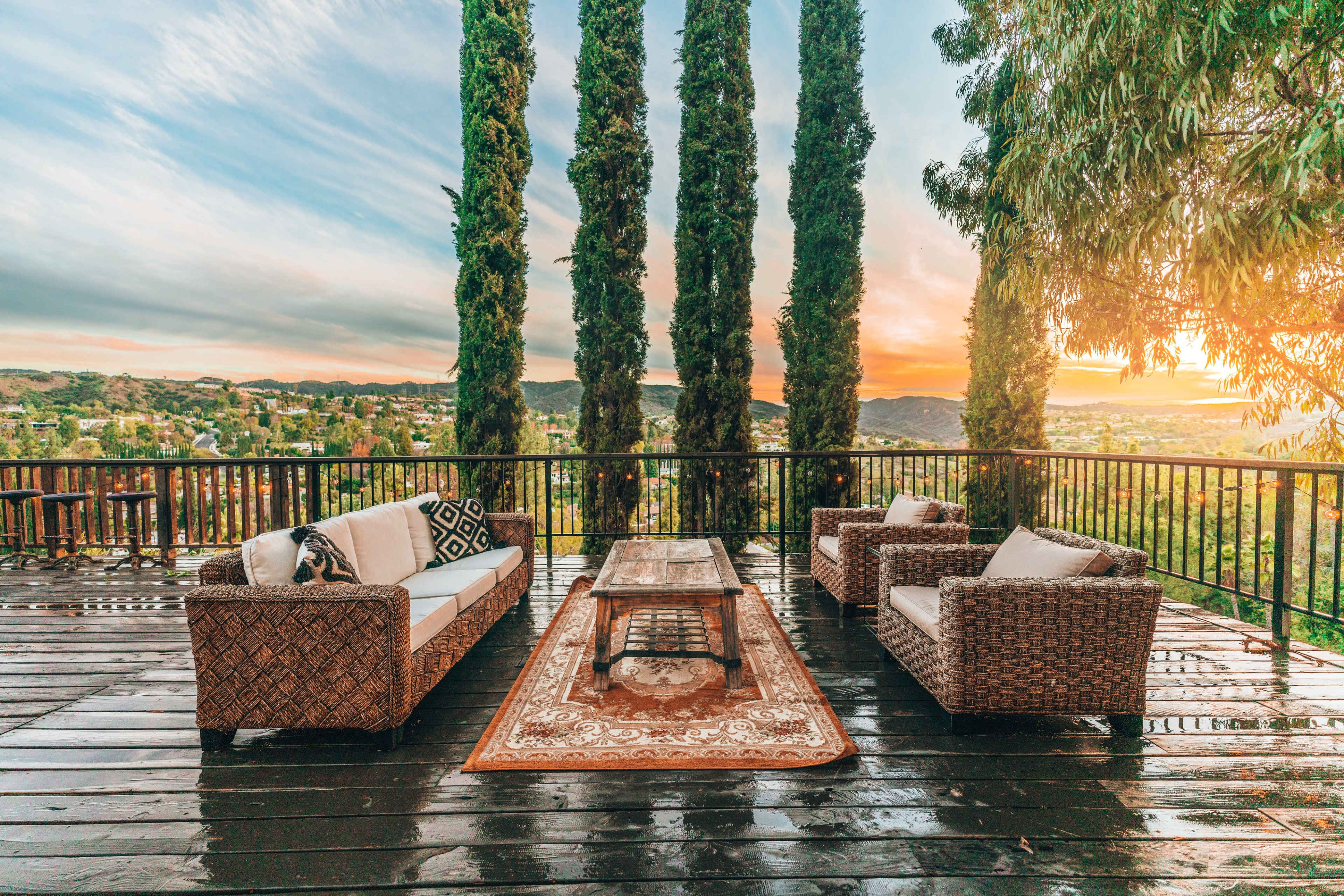 A wooden deck features two woven seating areas with a central table, framed by tall trees and a sunset view over a valley.
