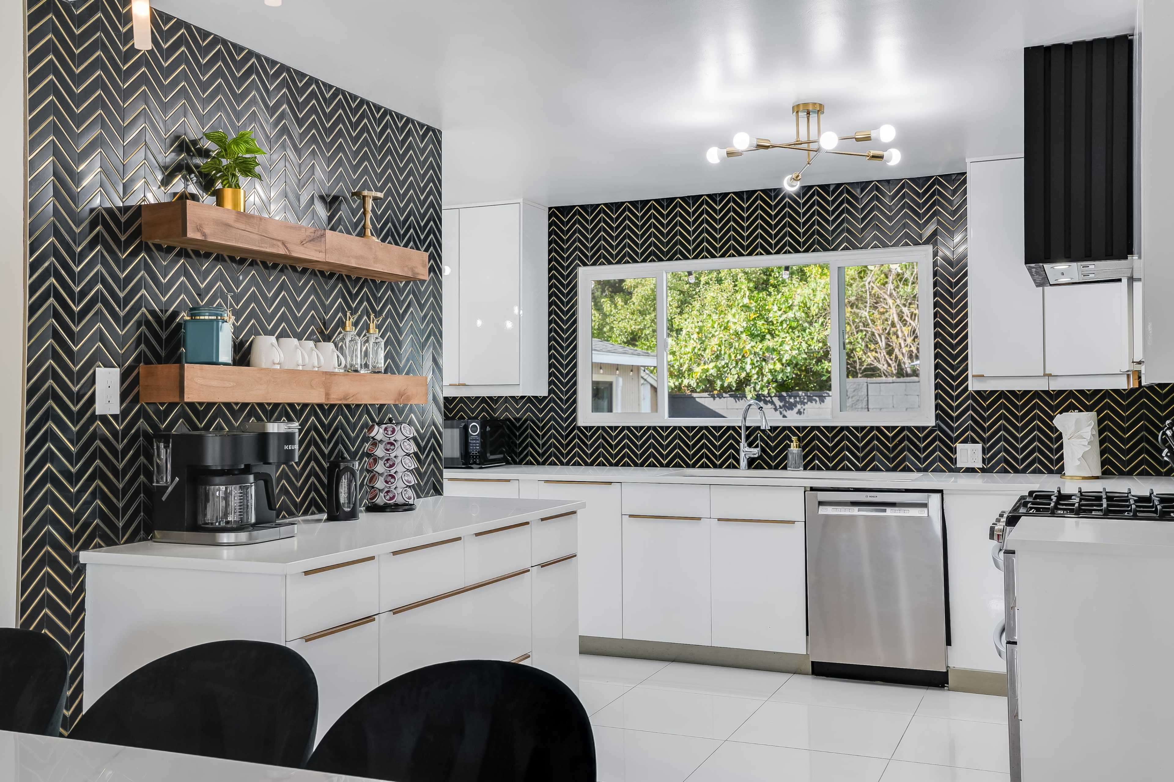 A modern kitchen features white cabinetry, black and gold patterned walls, wooden shelves, and a large window overlooking greenery.