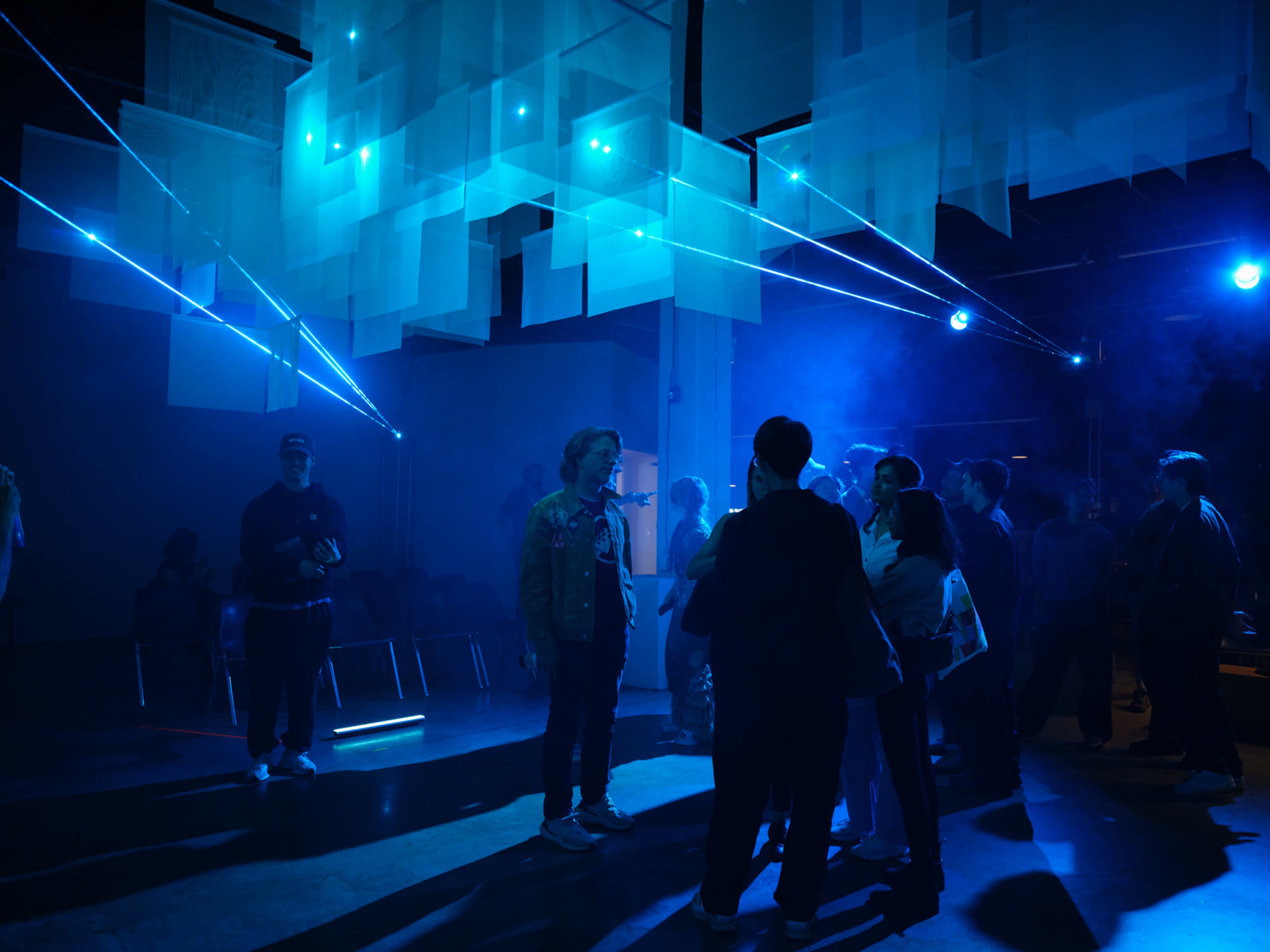 A group of people interact in a dimly lit space with blue laser beams cutting through translucent panels hanging from the ceiling.