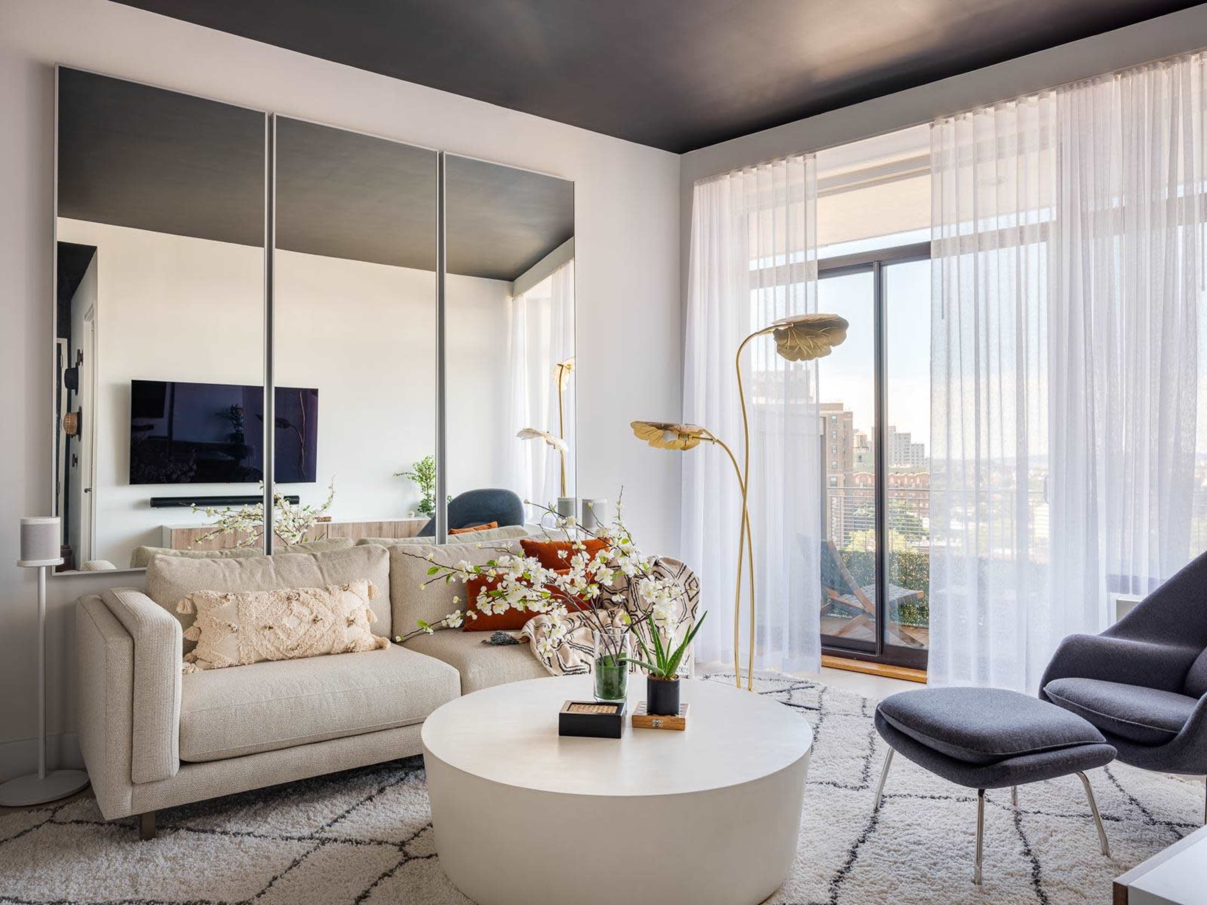 The image shows a modern living room with a light-colored sofa, a round coffee table, and large windows covered by sheer curtains, revealing a city view.