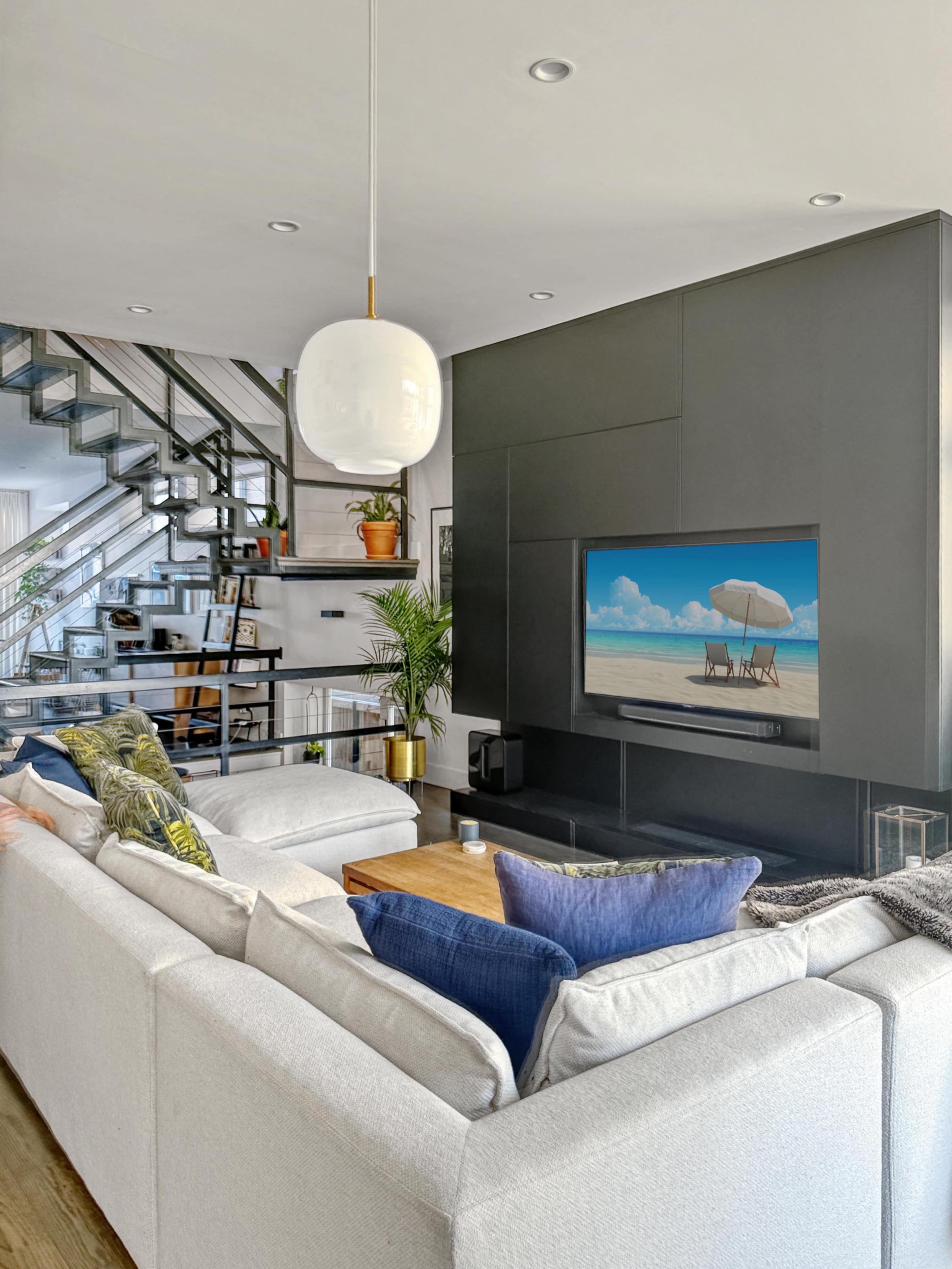 A modern living room features a large sectional sofa facing a flat-screen TV mounted on a dark wall, with a staircase visible in the background.