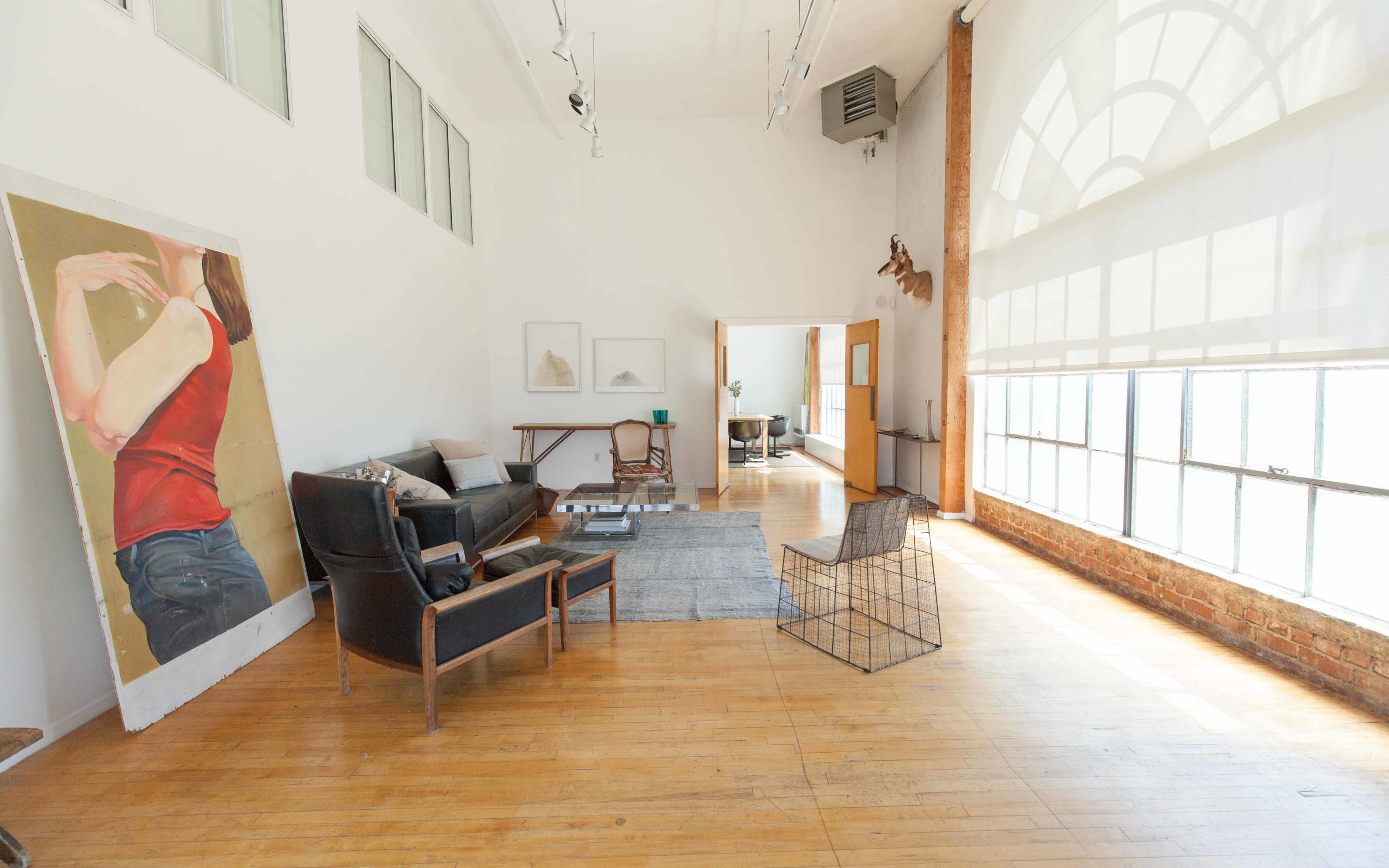 The image shows a spacious, well-lit loft-style room with hardwood floors and minimalistic furniture, including a sofa, a wire chair, and large windows.