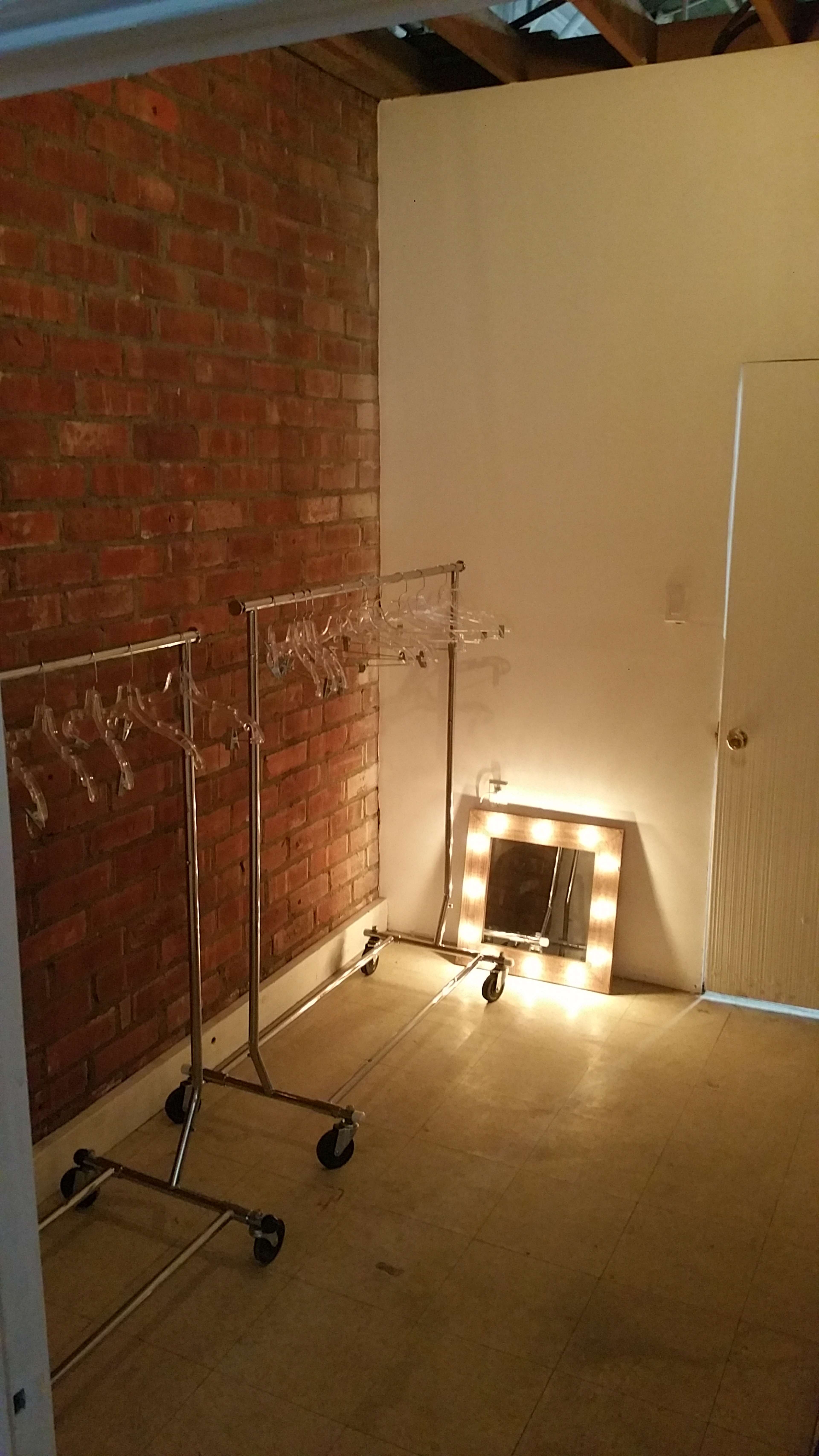 The image shows an empty room with exposed brick walls, two clothing racks on wheels, and a mirrored vanity with light bulbs.