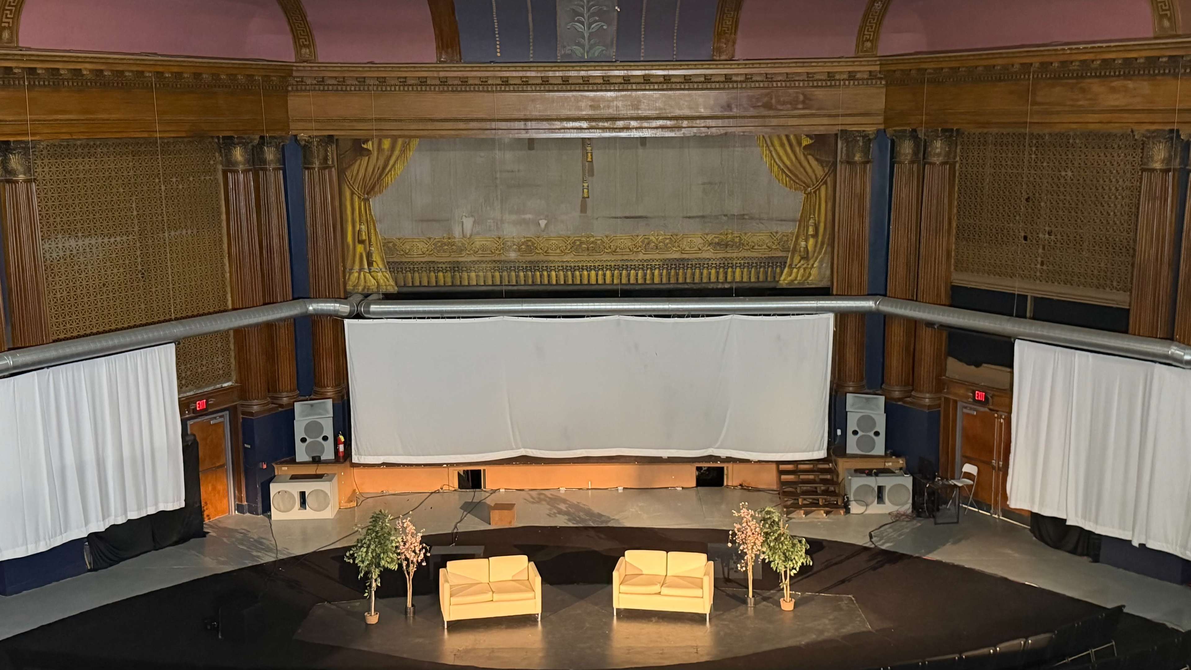 The image shows an empty theater stage with two chairs and decorative plants, framed by large curtains and flanked by sound equipment.