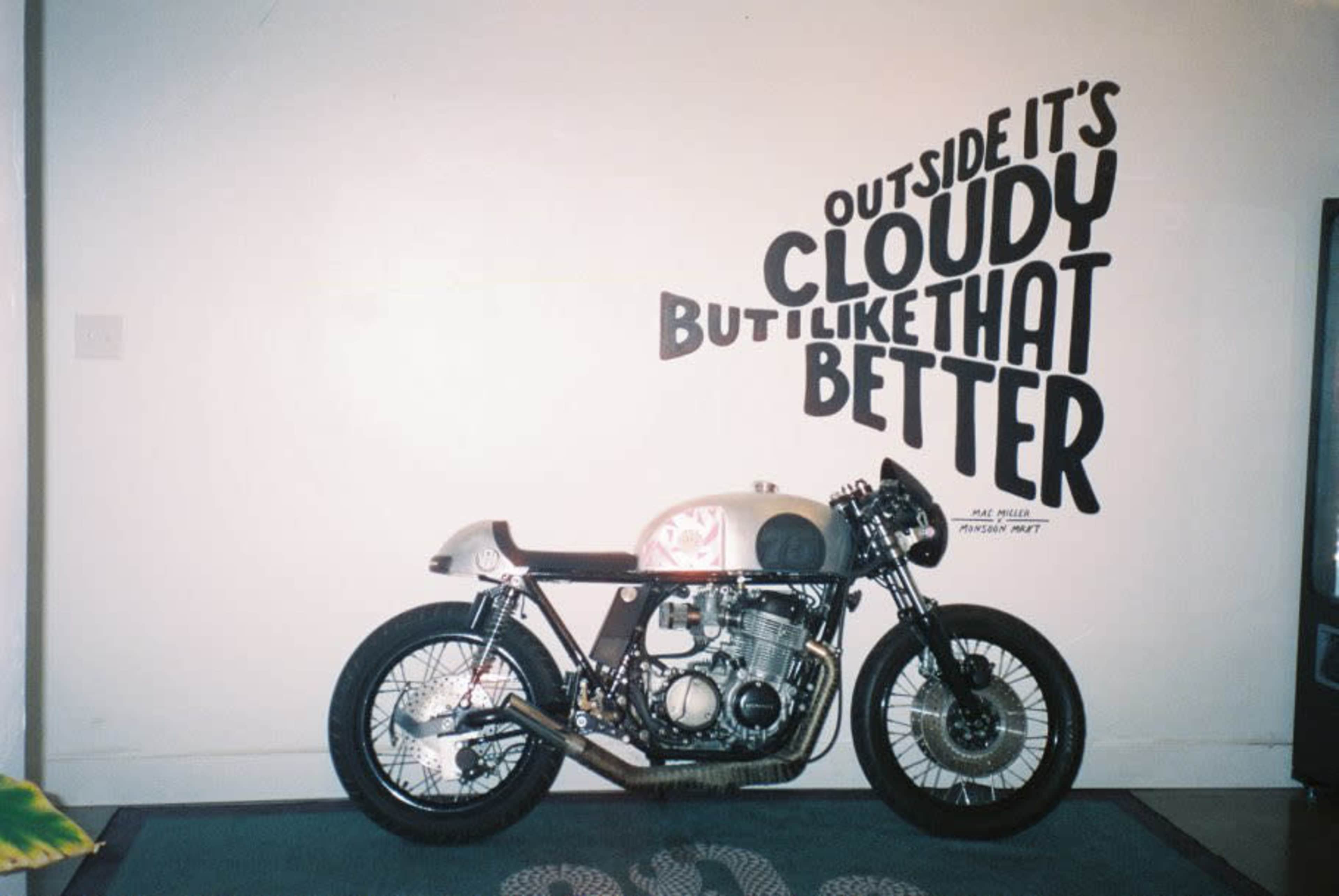 A motorcycle is positioned against a wall featuring the text: "OUTSIDE IT'S CLOUDY BUT I LIKE THAT BETTER."