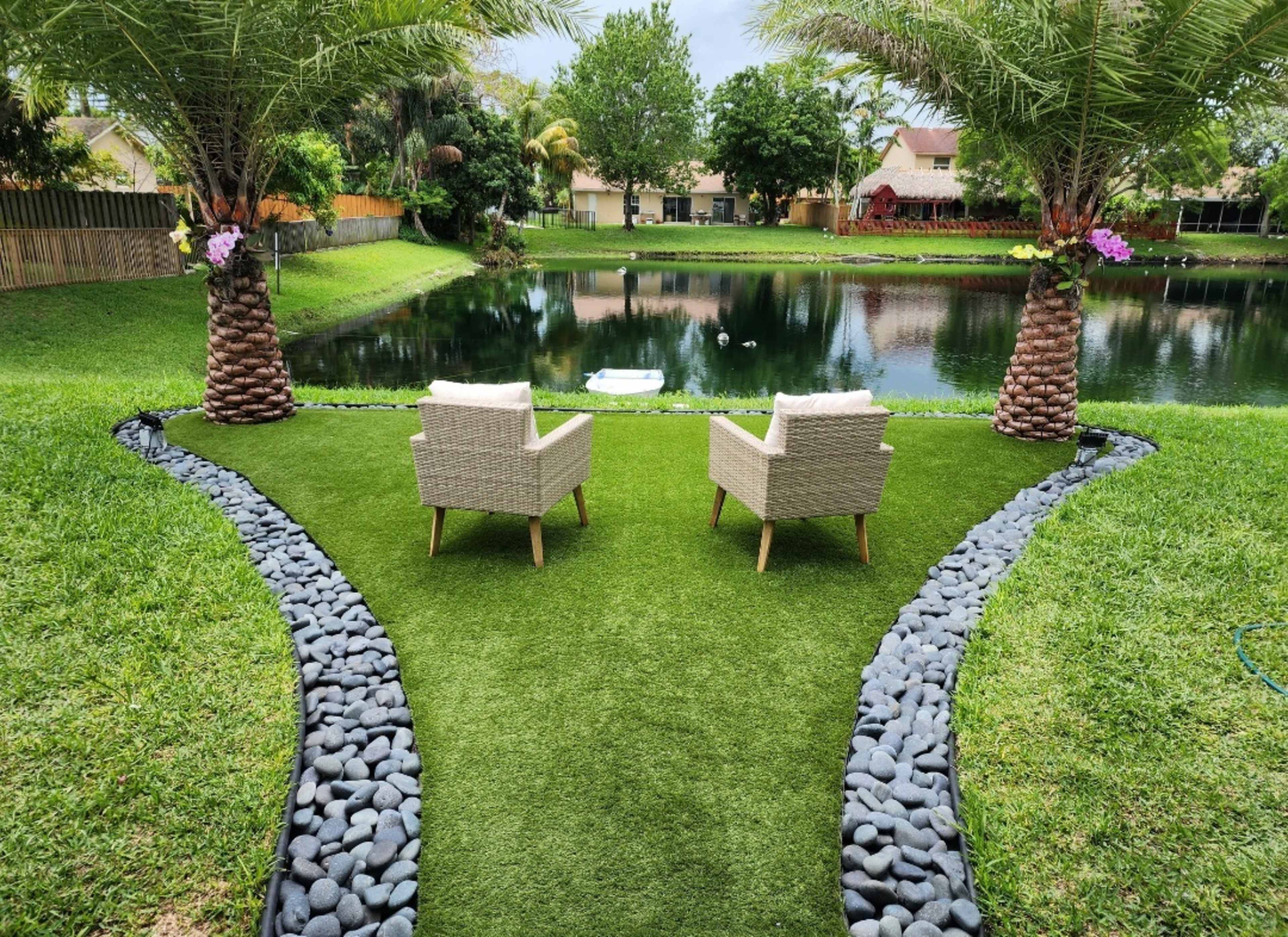 Two woven chairs are placed on a grass-like surface near a pond, flanked by palm trees and bordered by a stone path.