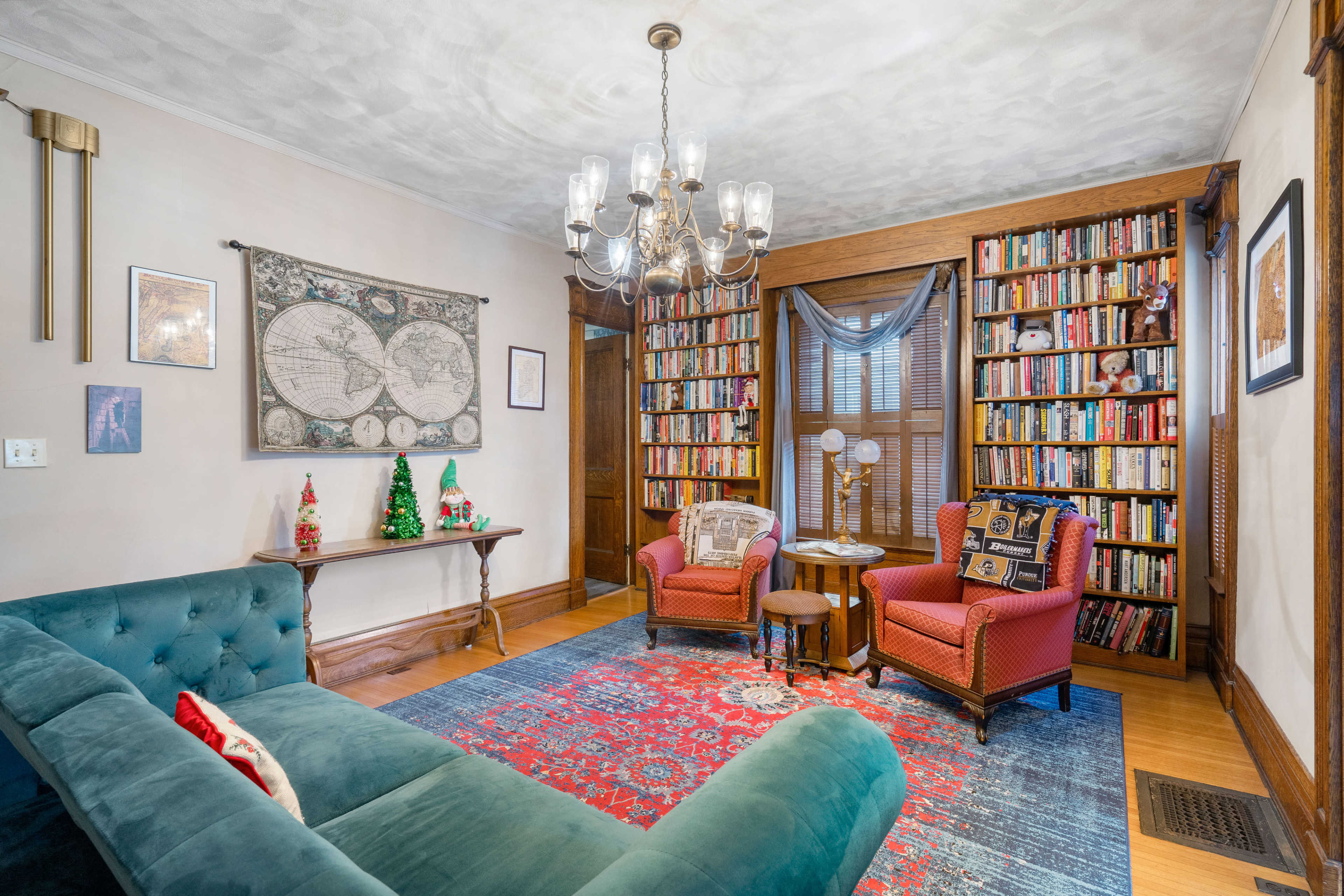 A cozy living room featuring a blue sofa, two red armchairs, a wooden bookshelf filled with books, and a chandelier overhead.