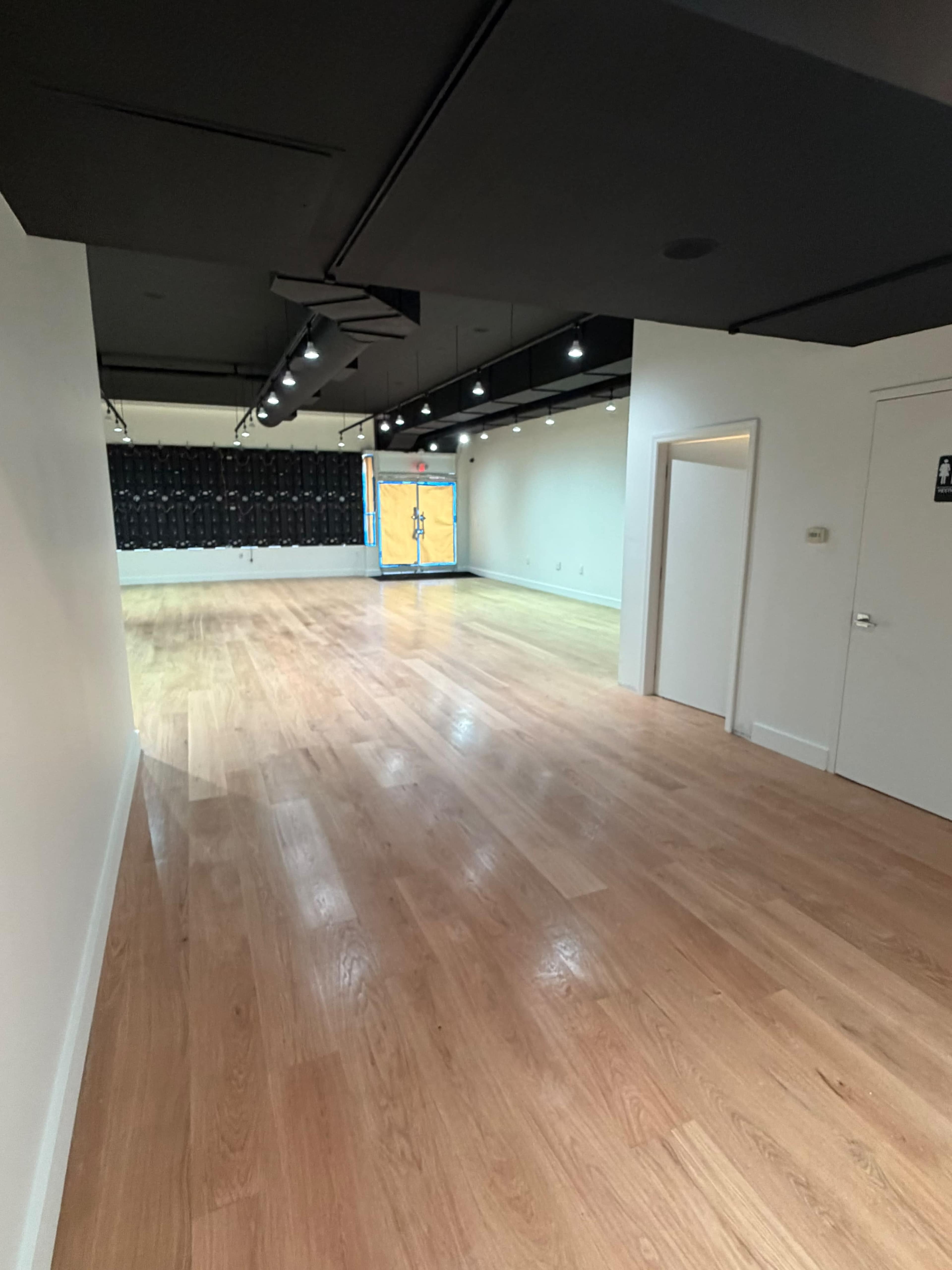The image shows a spacious, empty room with wooden flooring and bright overhead lights, featuring a door on one side and a wall with a dark backdrop at the far end.