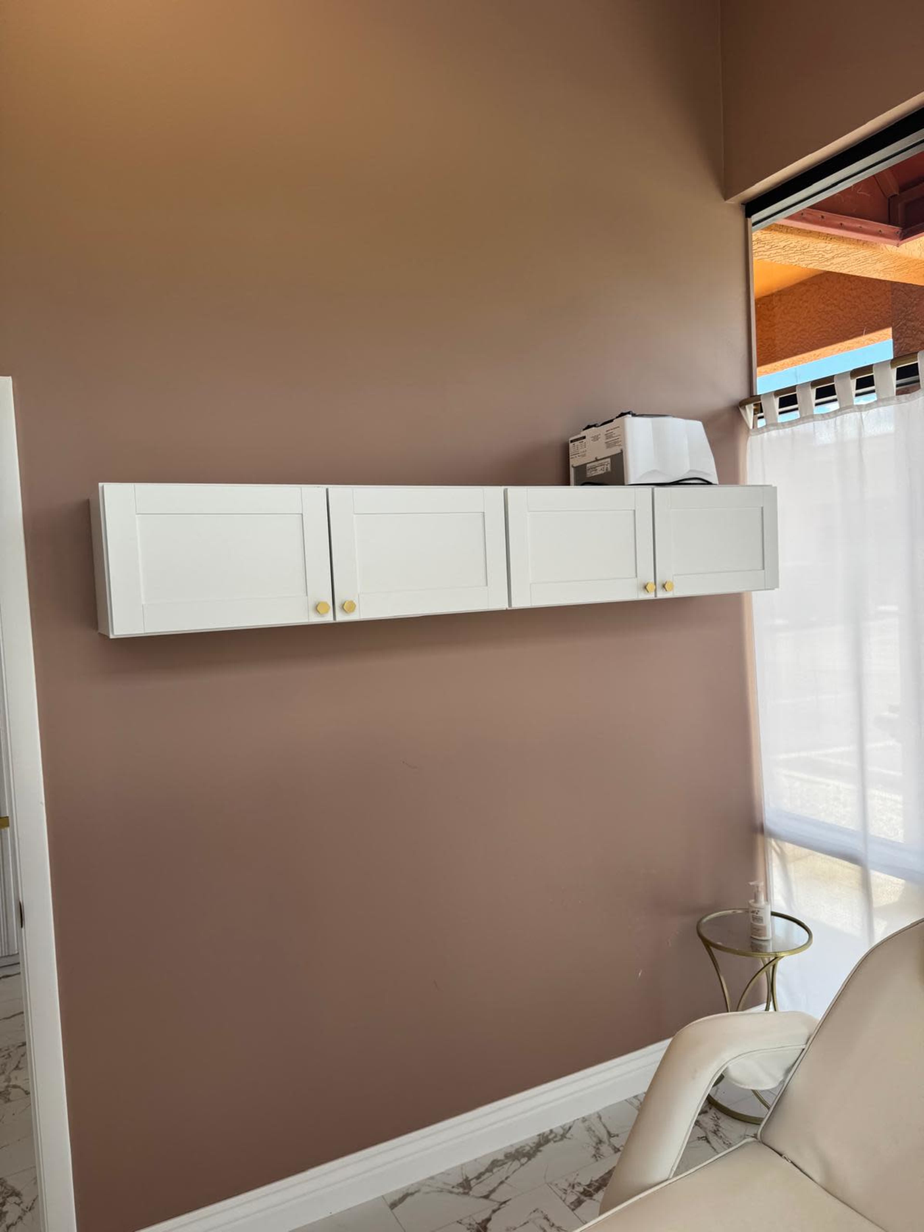 The image shows a white wall-mounted cabinet with three doors set against a brown wall, with a window and a white chair visible nearby.