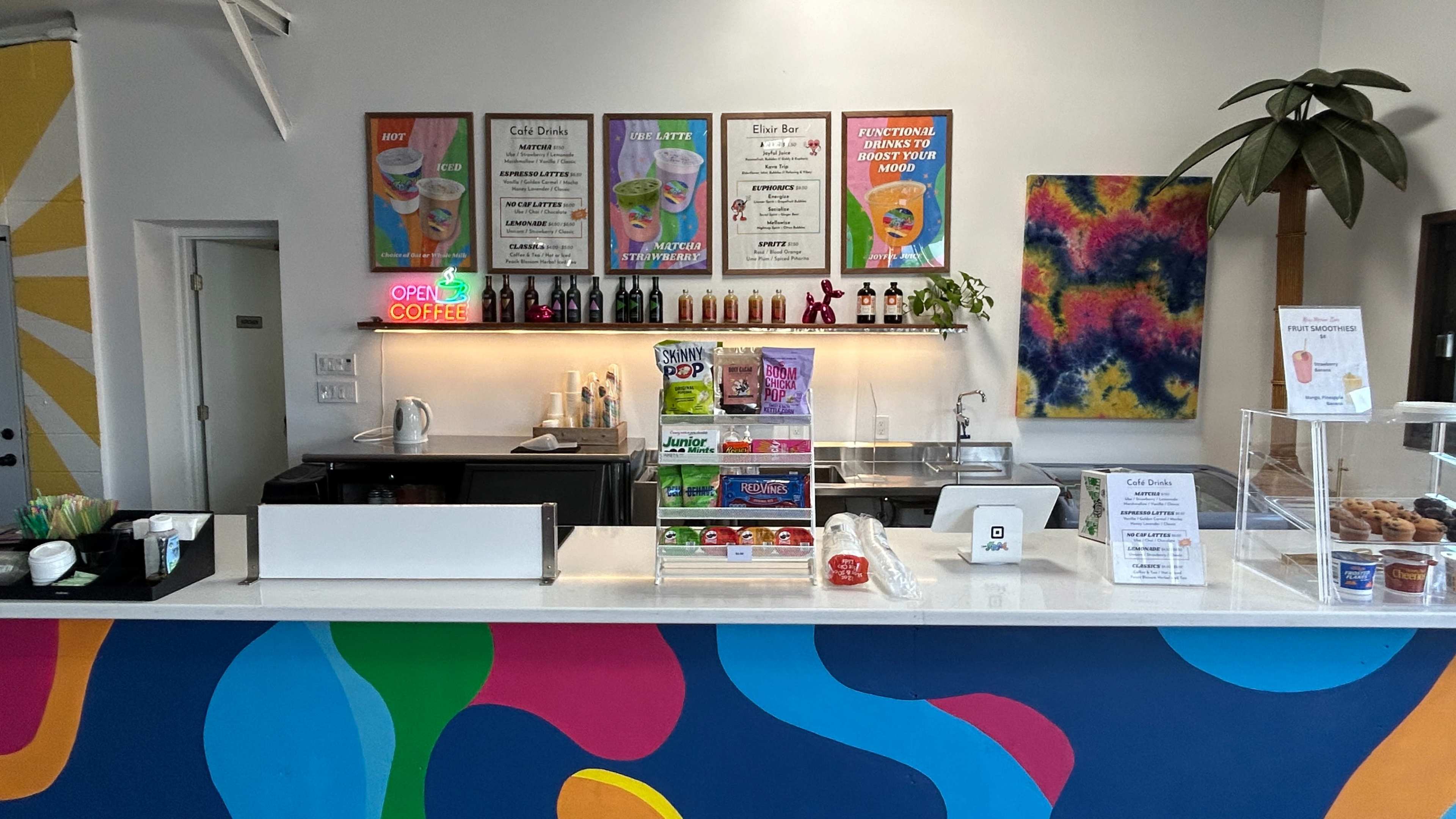 The image shows a colorful café counter with a menu display, beverages, and decorative artwork on the walls.