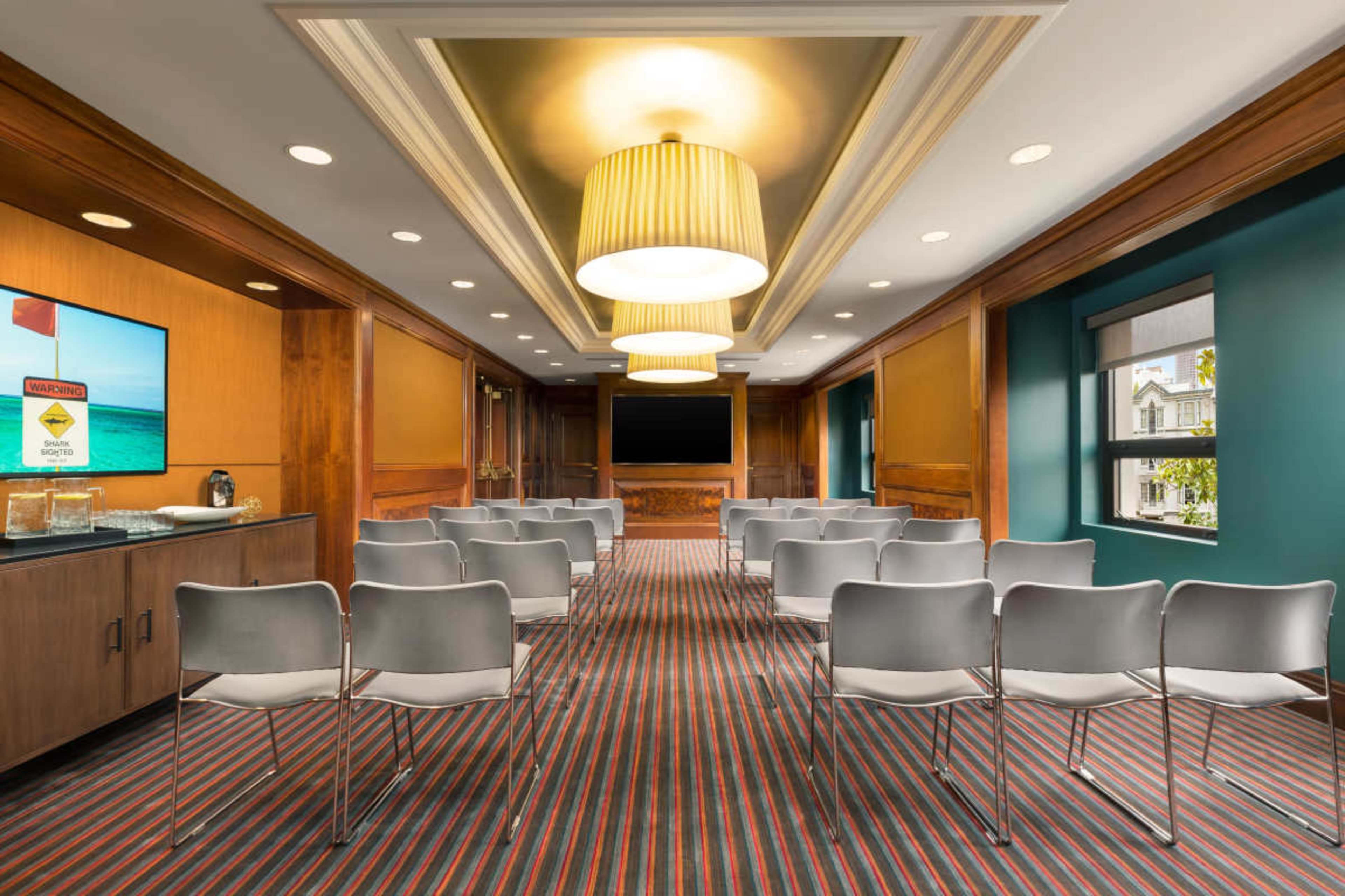 The image shows a conference room featuring a long central table, multiple rows of white chairs, and a large screen at the front, with warm-colored walls and modern lighting.