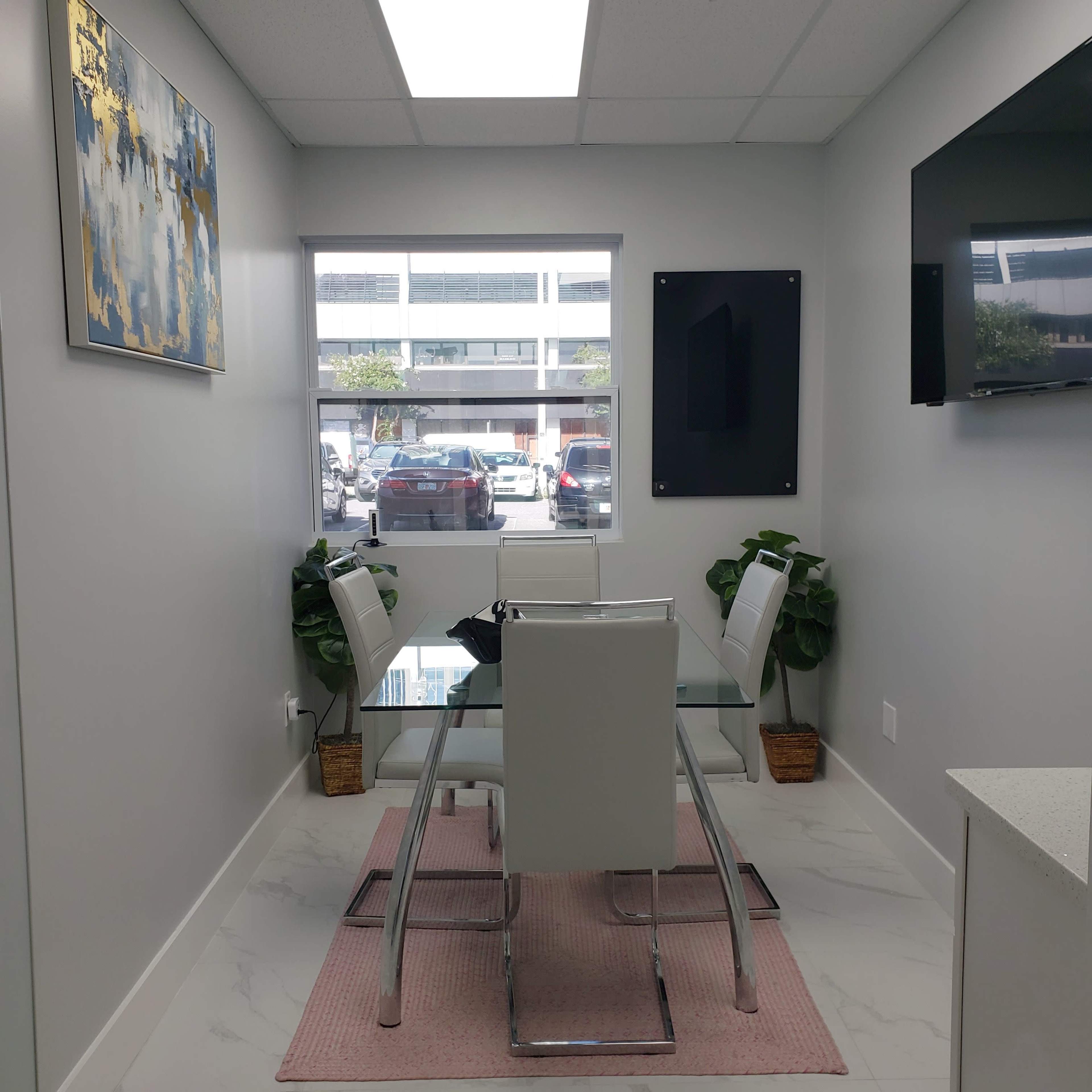 A small conference room with a glass table surrounded by four white chairs, and a window overlooking a parking lot.