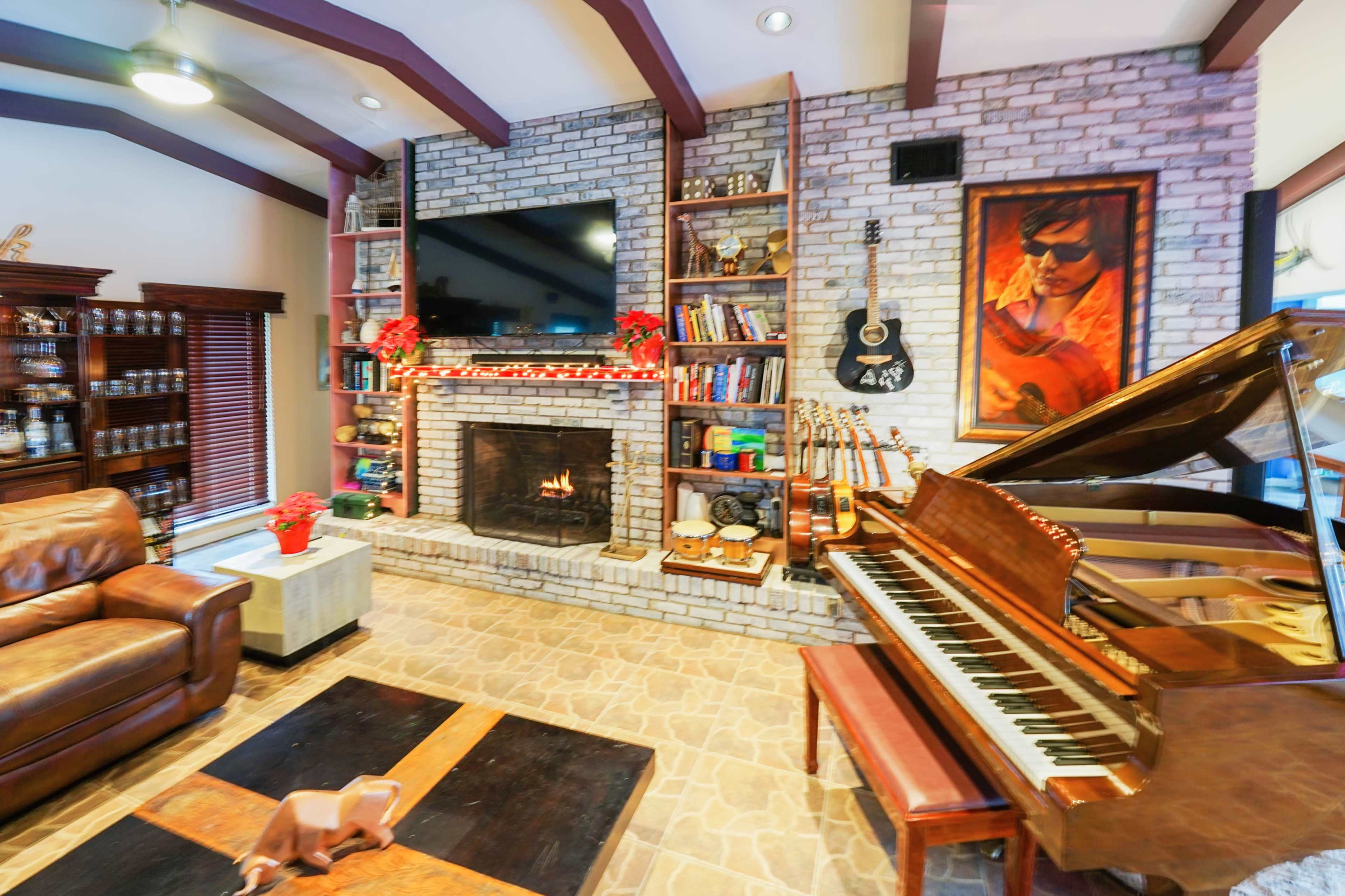 The image shows a cozy living room with a brick fireplace, a piano, bookshelves filled with books and decorations, and a leather couch.