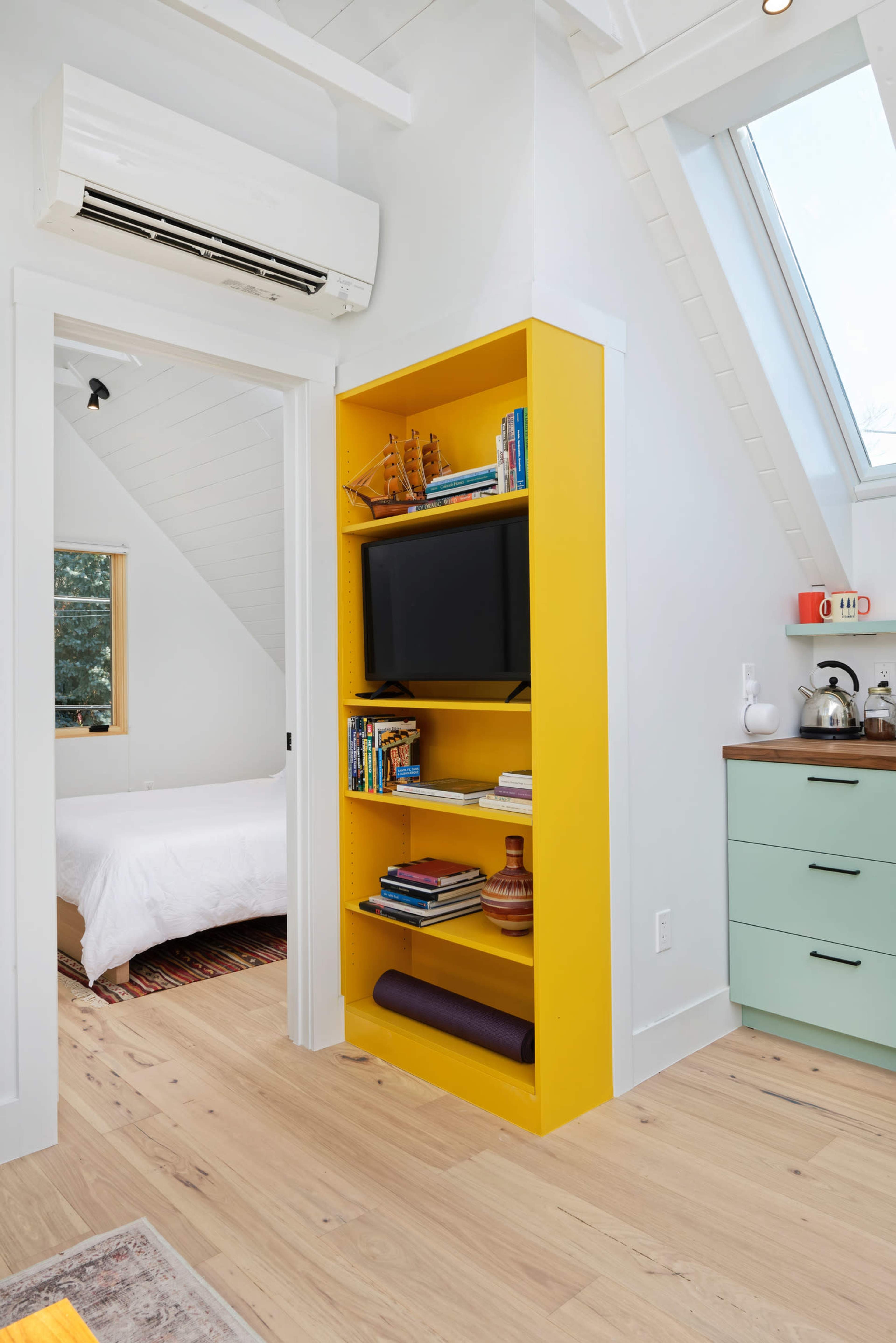 A bright yellow bookshelf stands next to a doorway in a modern room, featuring a television and several books, with a bed visible in the background.