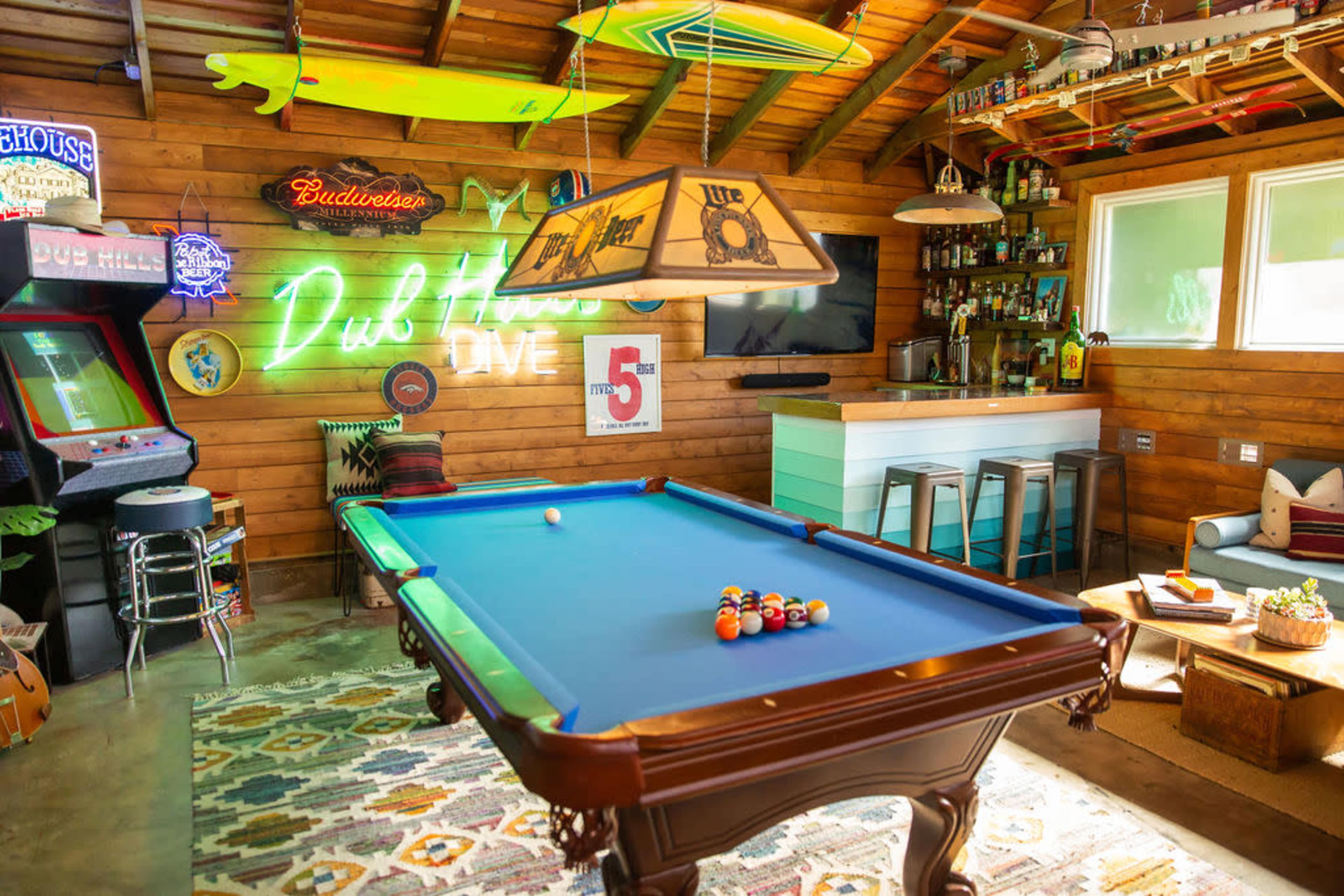 A cozy game room featuring a blue pool table, a vintage arcade game, and a bar area with neon signs and surfboards hanging on the walls.