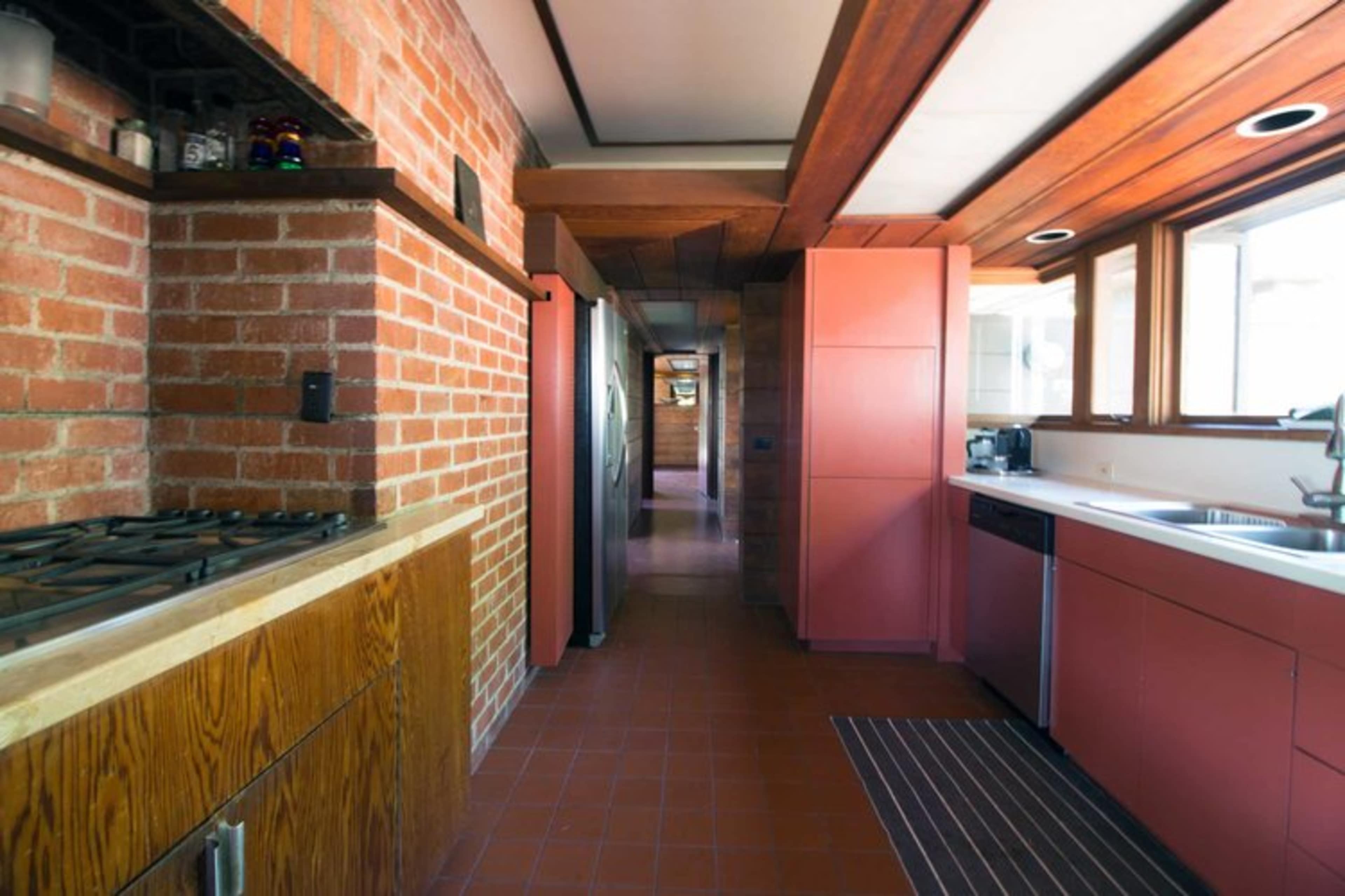 The image shows a kitchen featuring brick walls, wooden beams, and pink cabinets, with a visible corridor leading to the back of the room.