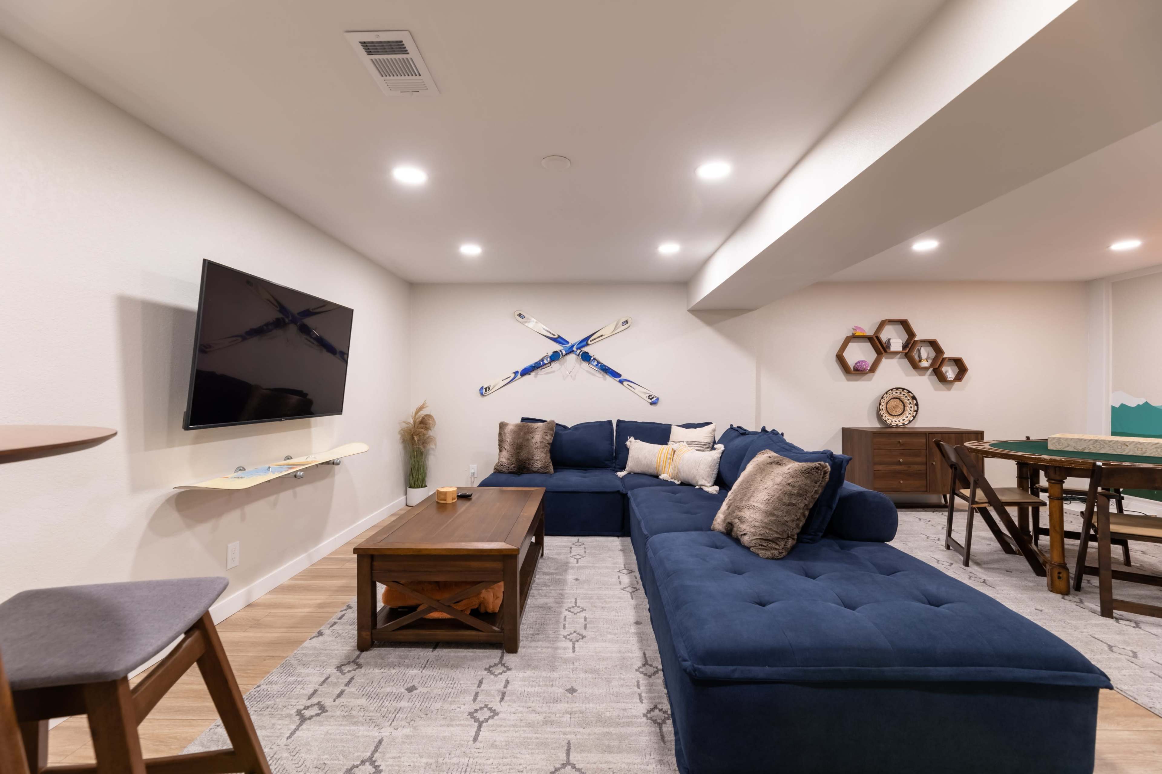 A modern basement living area with a dark blue sectional sofa, a wooden coffee table, and decorative wall accents including ski equipment and hexagonal shelves.