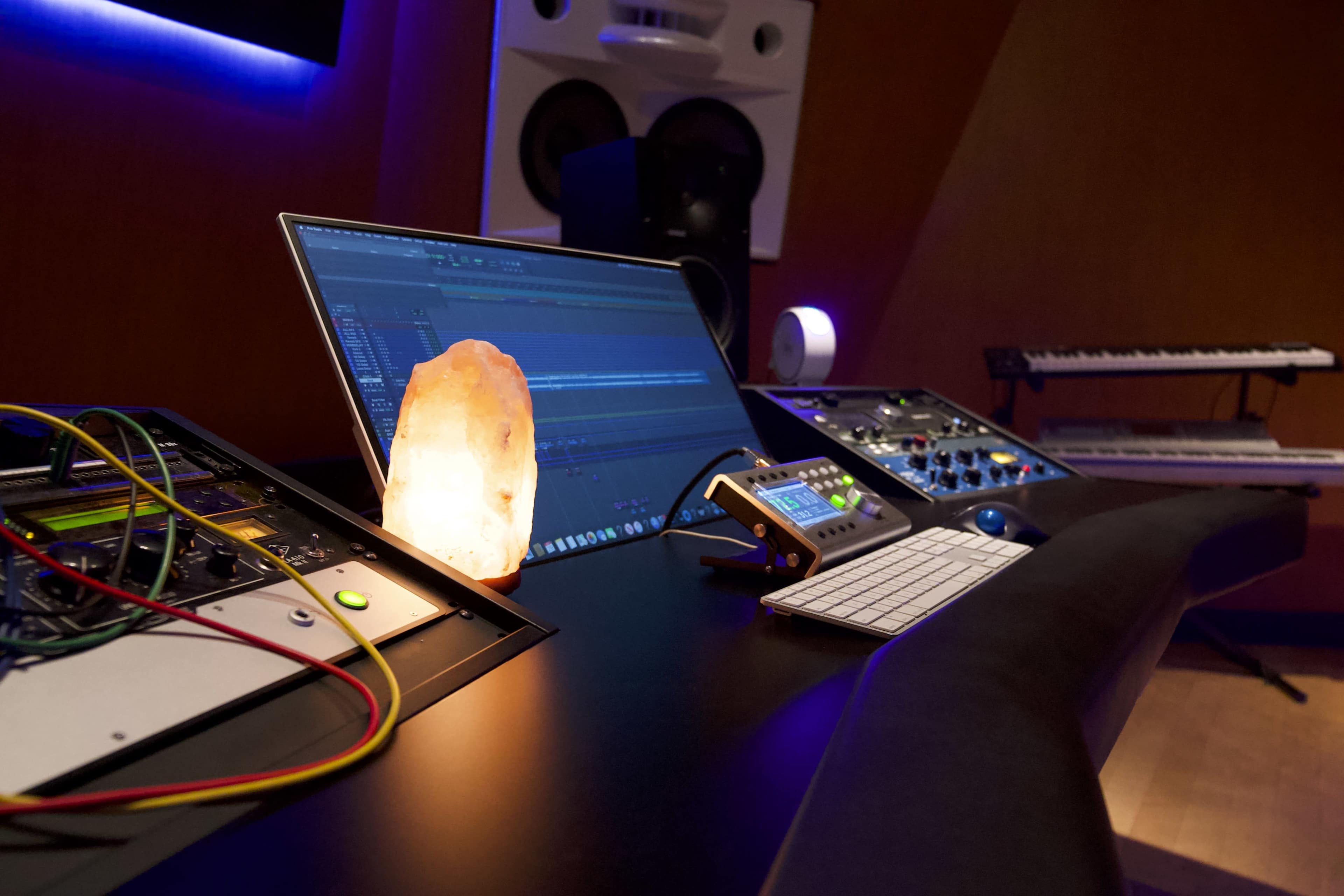 A modern music production studio featuring a computer monitor, mixing console, keyboard, and a salt lamp on a sleek desk.