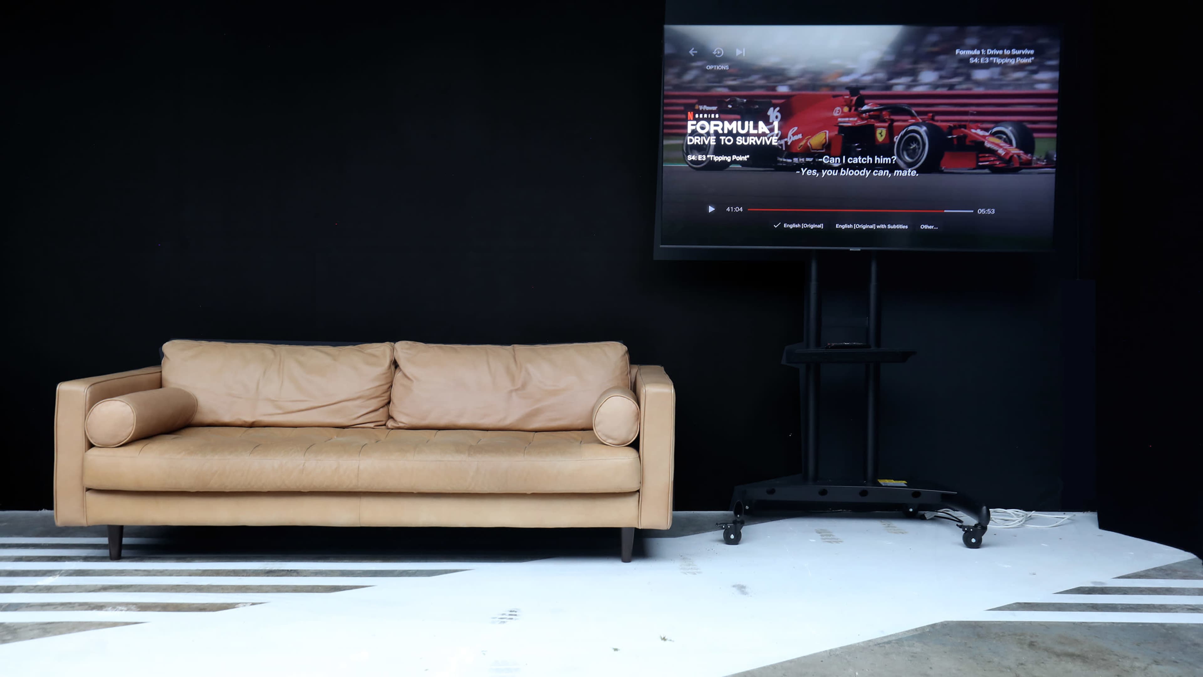 A tan leather couch is positioned in front of a large TV screen displaying a Formula 1 racing program in a minimalist black and white room.