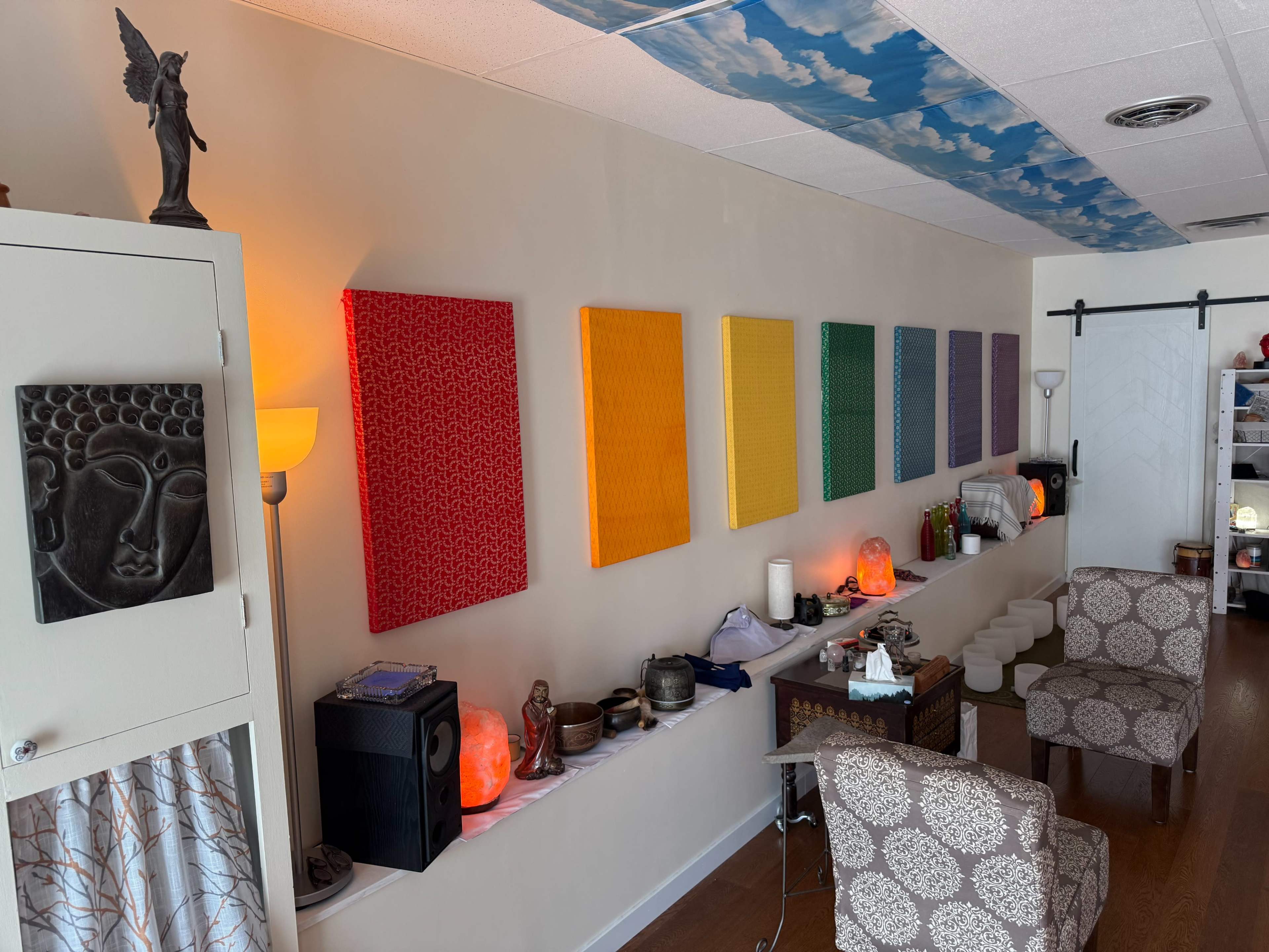 The image shows a serene interior space featuring a row of colorful canvases on the wall, a statue, various decorative items, and lamps.