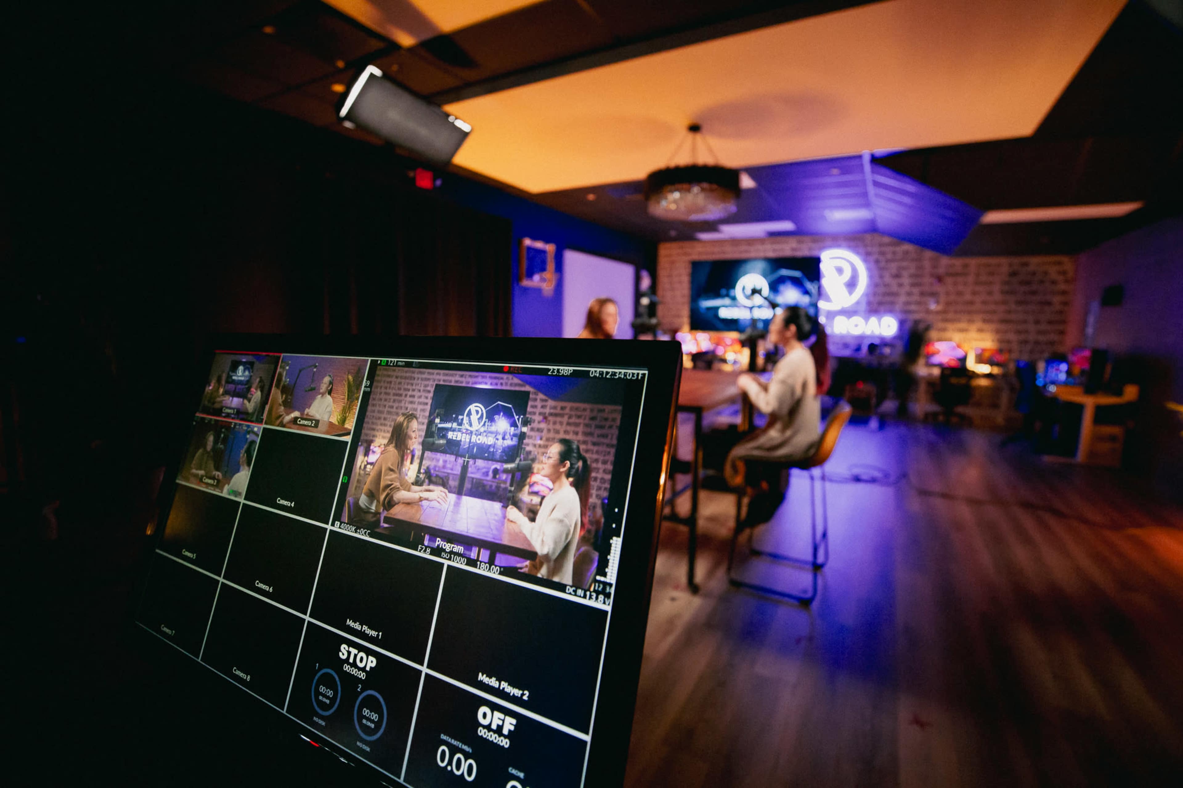 A studio setup for a video production, featuring a control monitor displaying camera angles while two people engage in conversation at a table in the background.