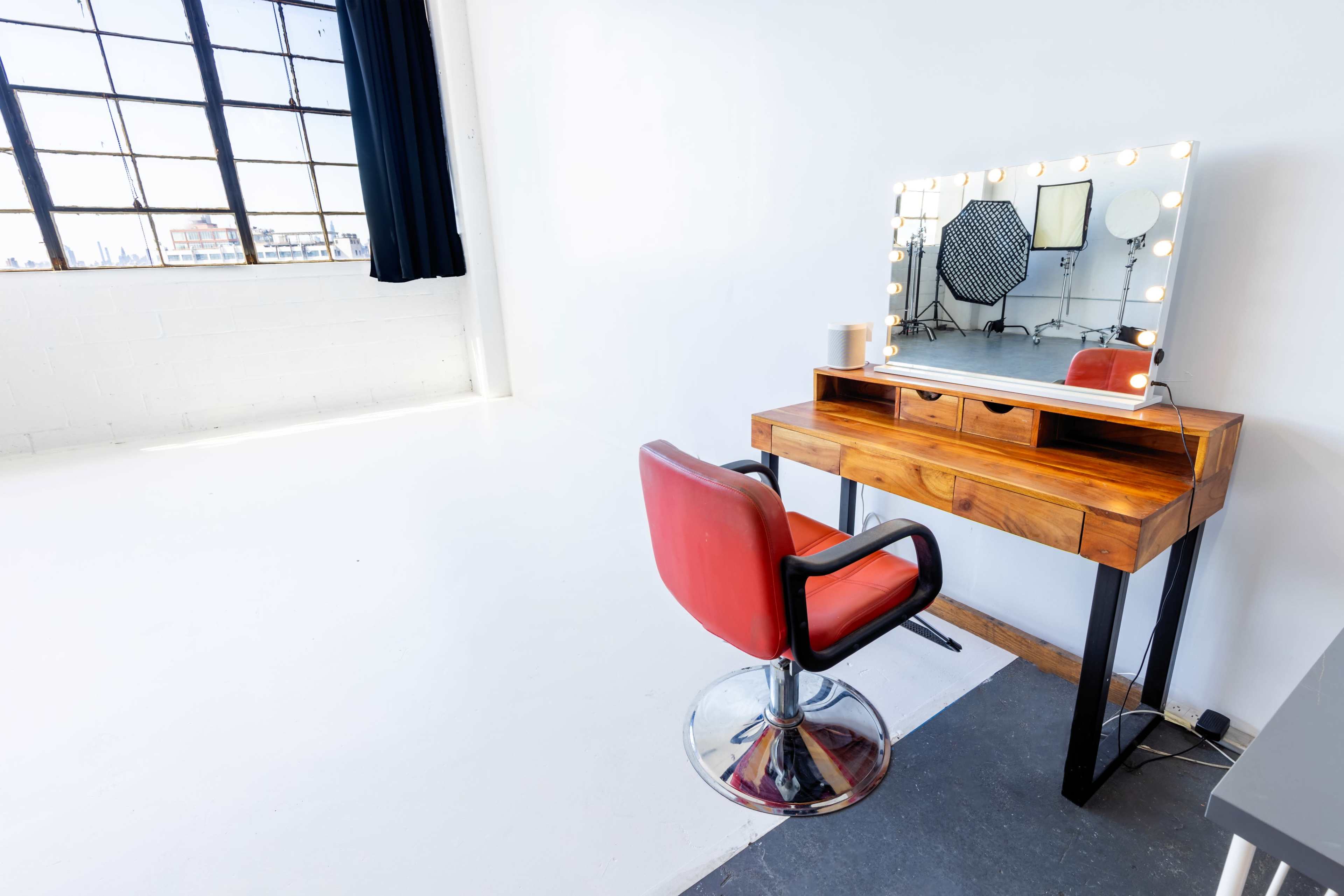 A modern hairstyling station featuring a wooden vanity with a mirror illuminated by bulbs and a red, retro-style chair, set against a bright, minimalist studio backdrop.