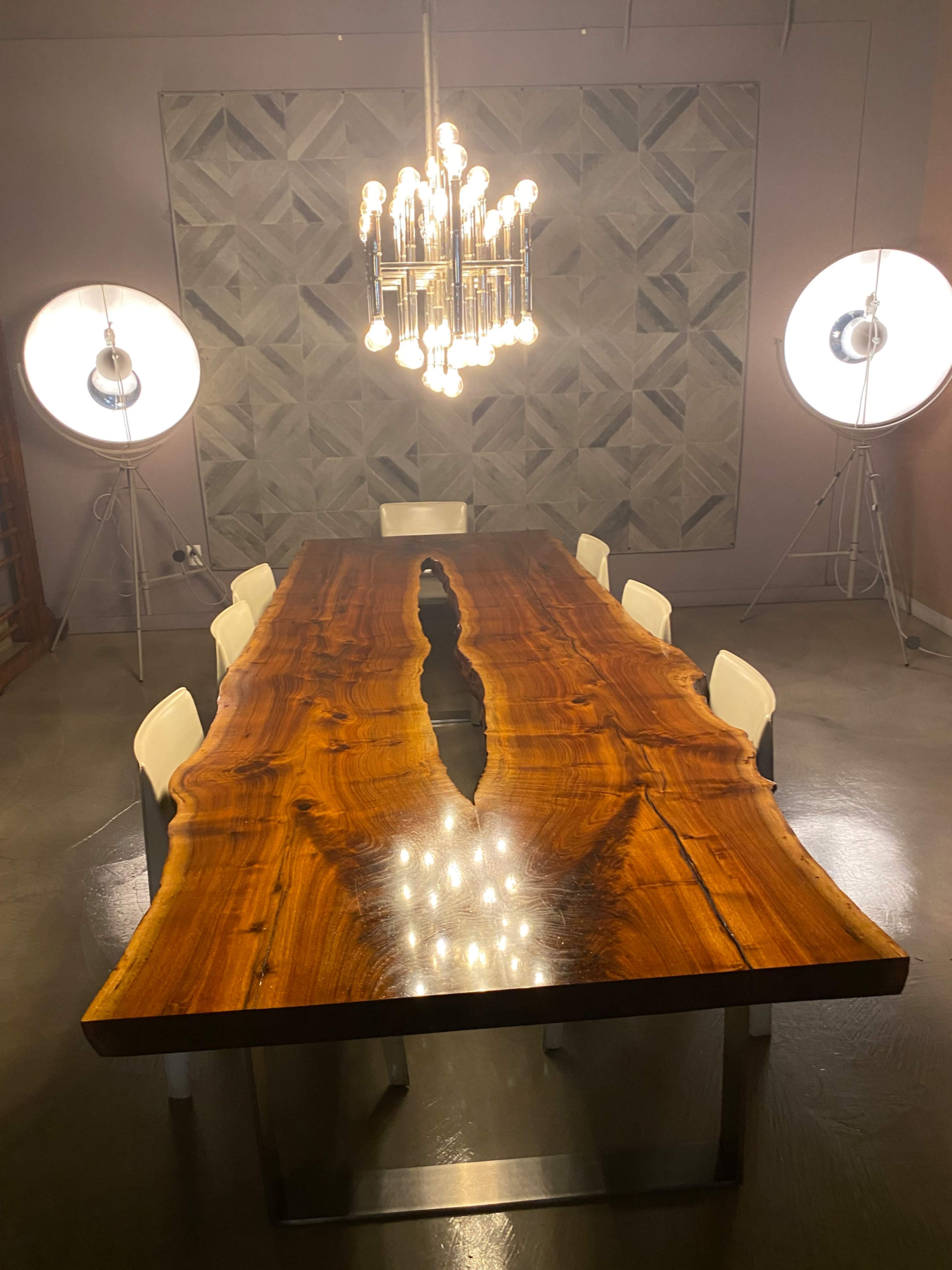 A large wooden dining table with a deep center crack is illuminated by a modern chandelier and flanked by six white chairs in a well-lit room.