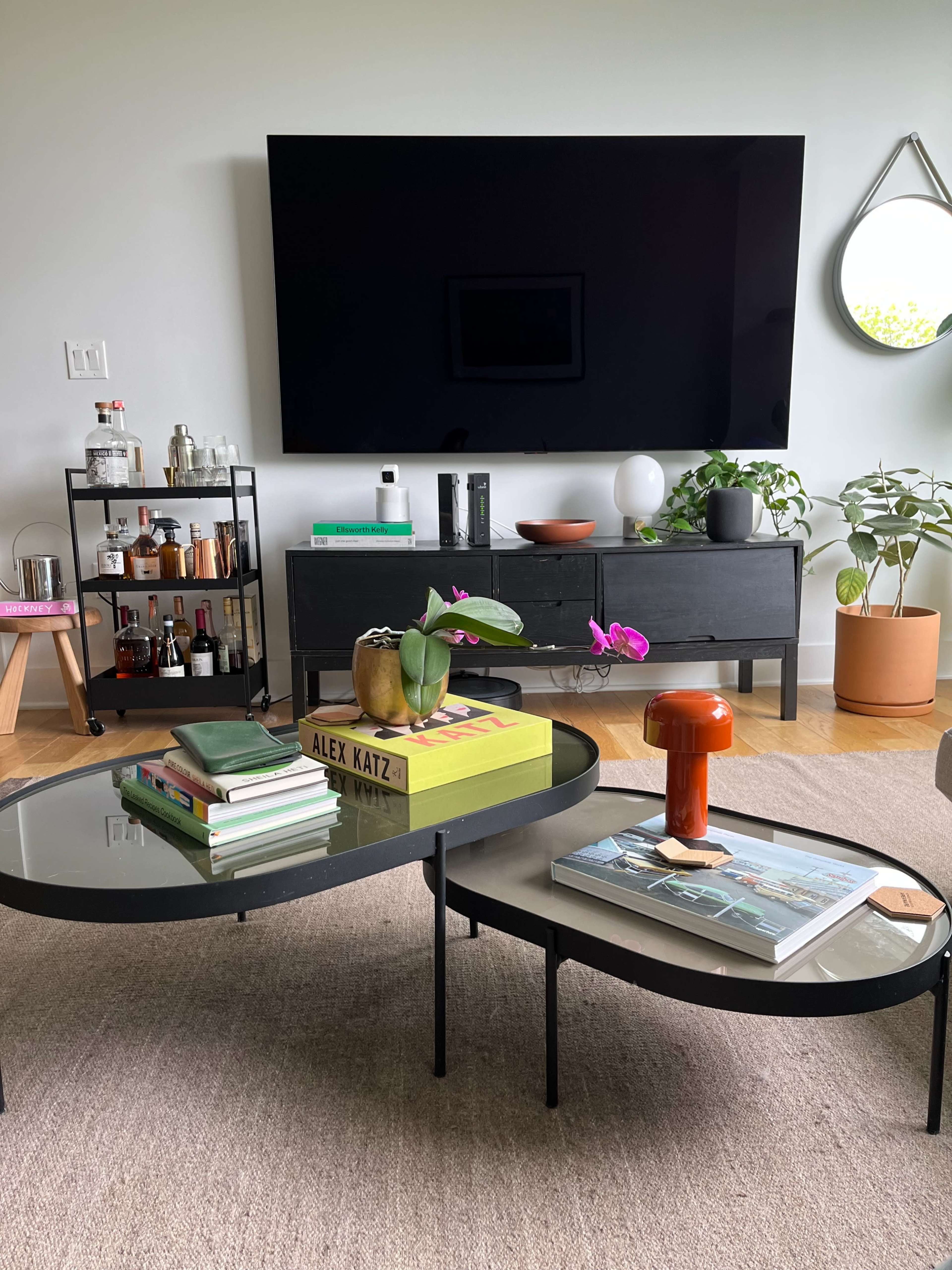 A modern living room with a large television mounted on the wall, two low coffee tables with books and decorative items, and a bar cart in the background.