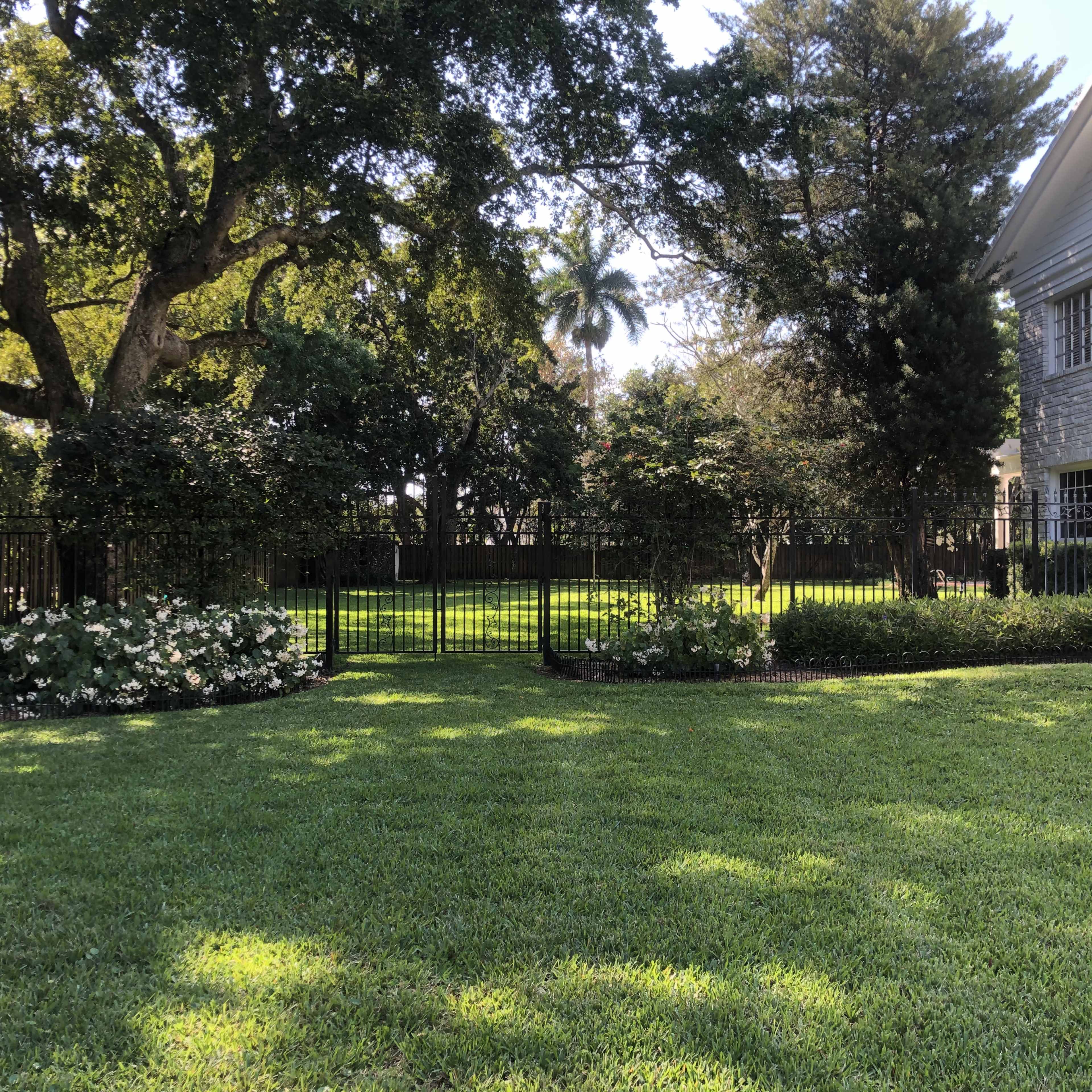 A well-maintained lawn with a black fence surrounds a garden featuring flowering plants and trees.