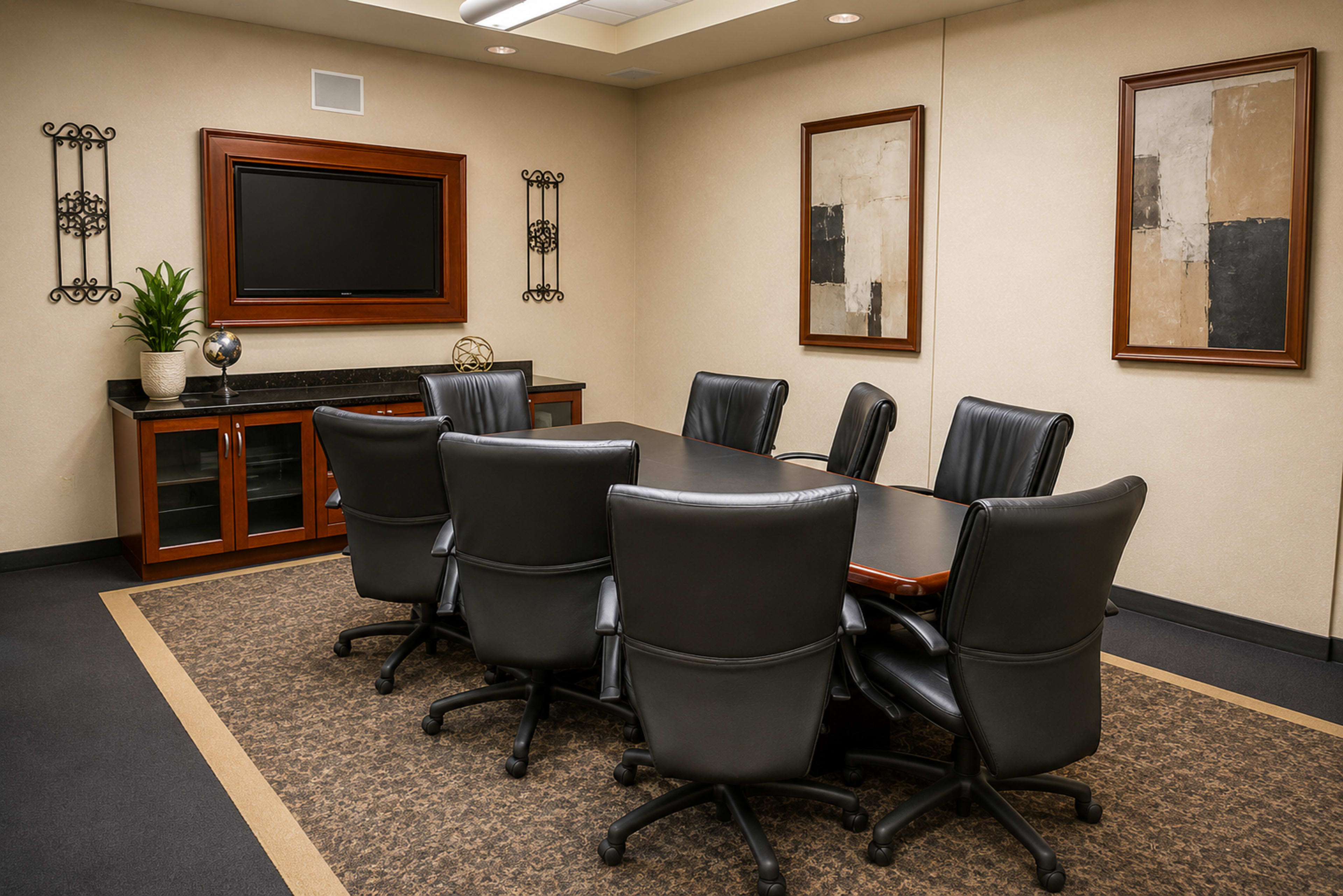 A modern conference room features a large table surrounded by black leather chairs, an entertainment unit with a TV mounted on the wall, and two framed abstract artworks.