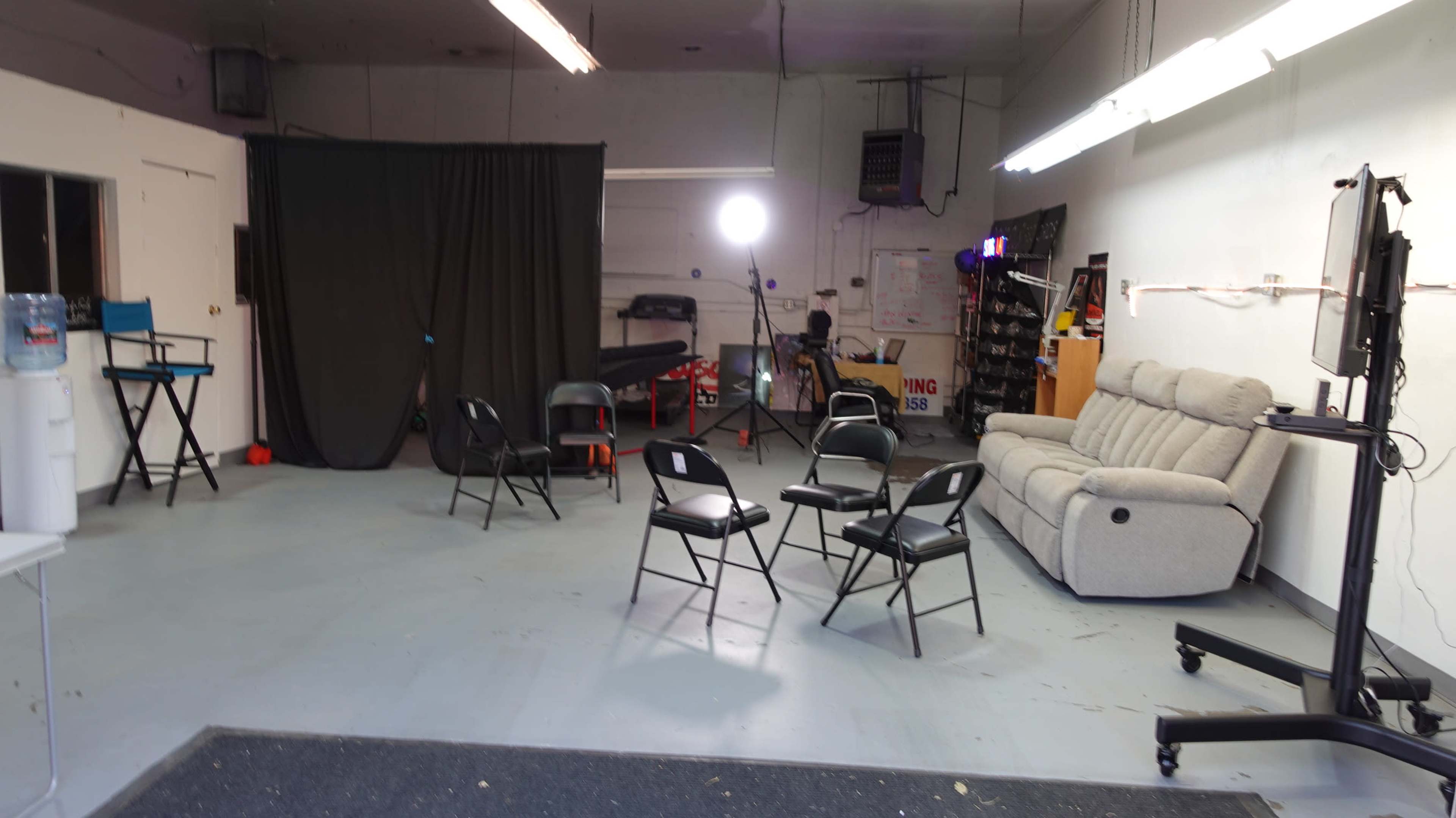 The image shows a spacious room with a couch, several black chairs arranged in a circle, and a light source pointed towards a dark curtain backdrop.