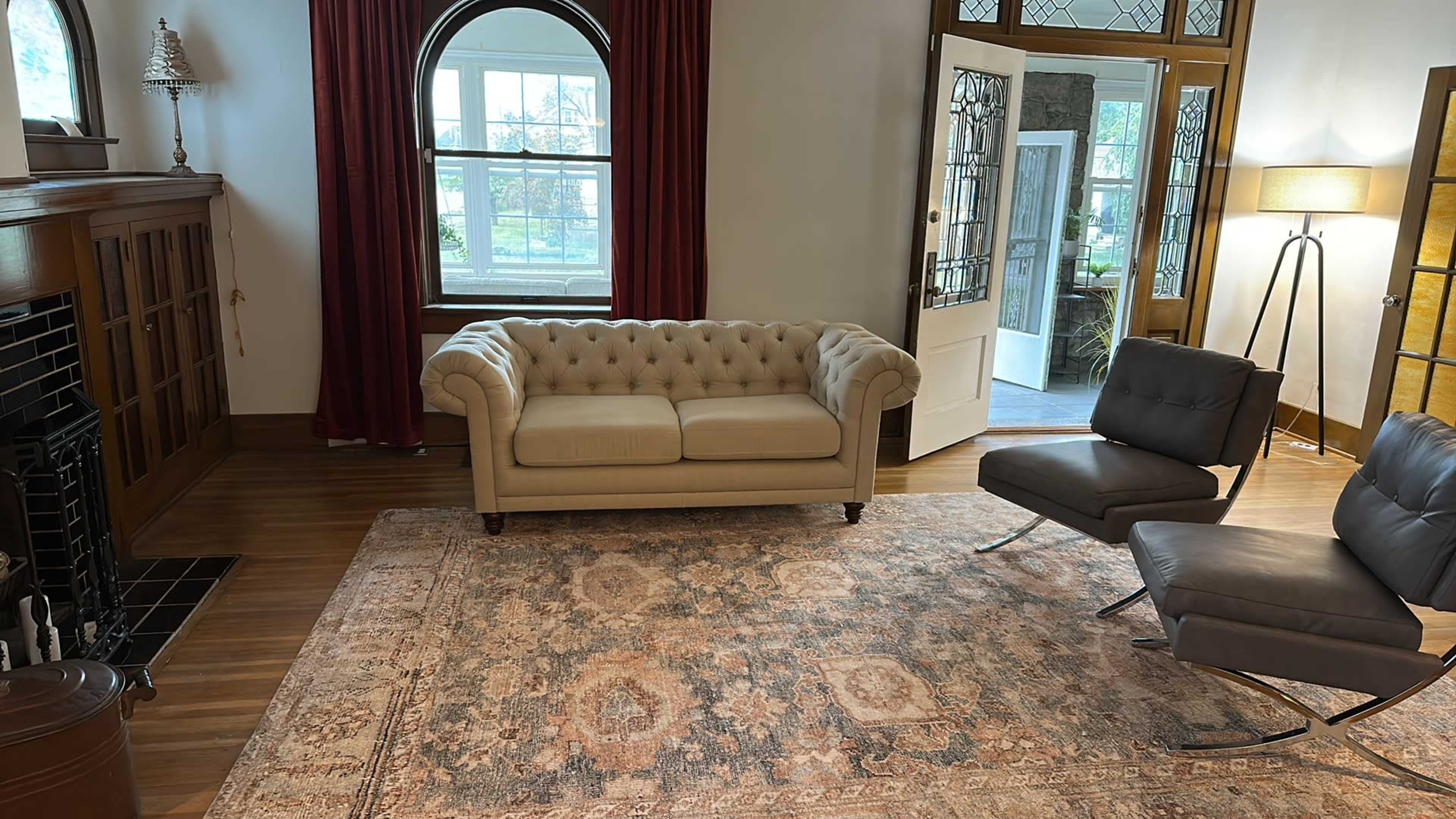 A beige tufted sofa and two dark gray chairs are arranged on an ornate area rug in a well-lit room with large windows and red drapes.