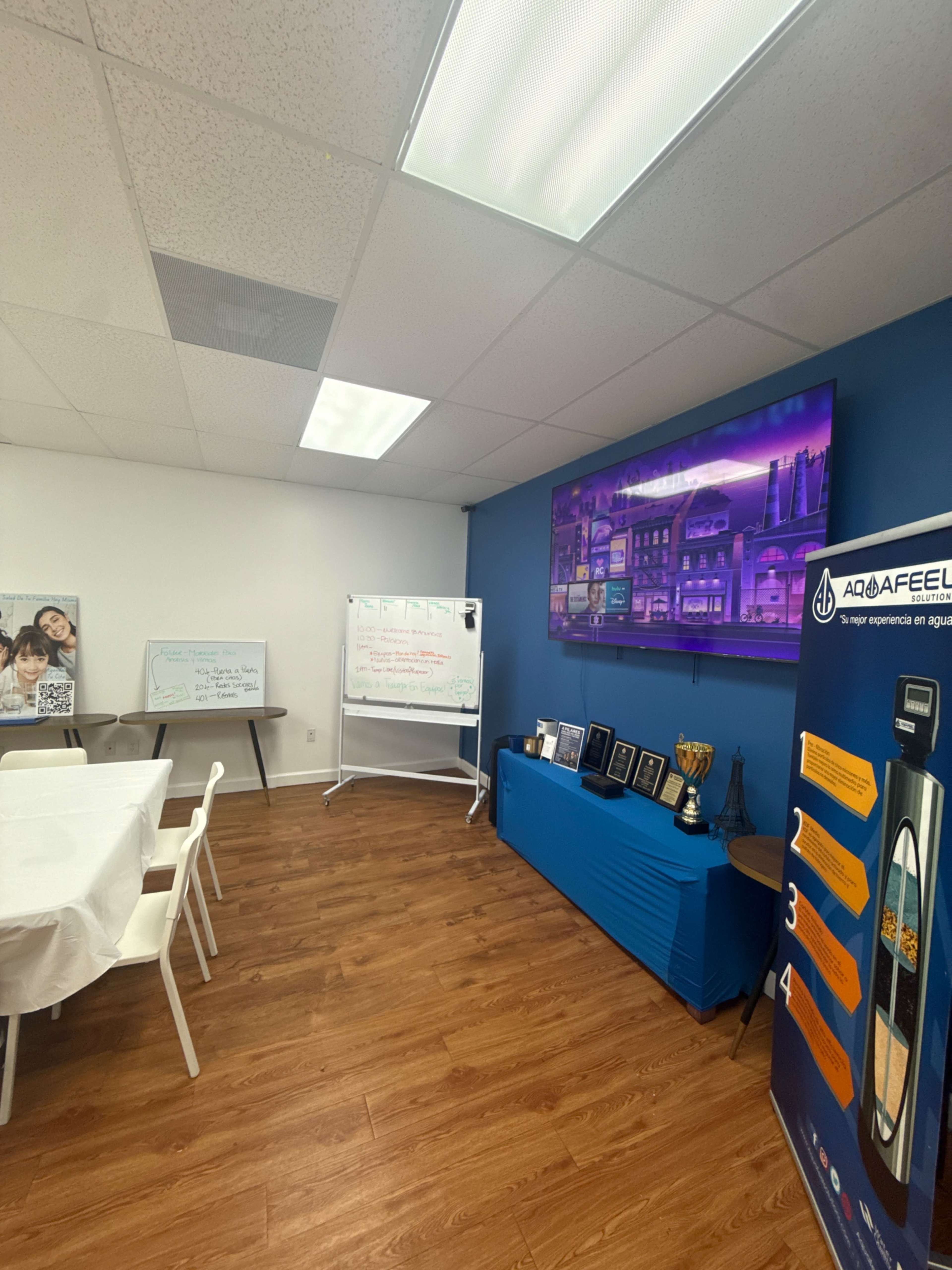 The image shows a meeting room with a large screen displaying graphics, a whiteboard, a table covered with a cloth, several chairs, and a blue backdrop featuring a logo and trophies.