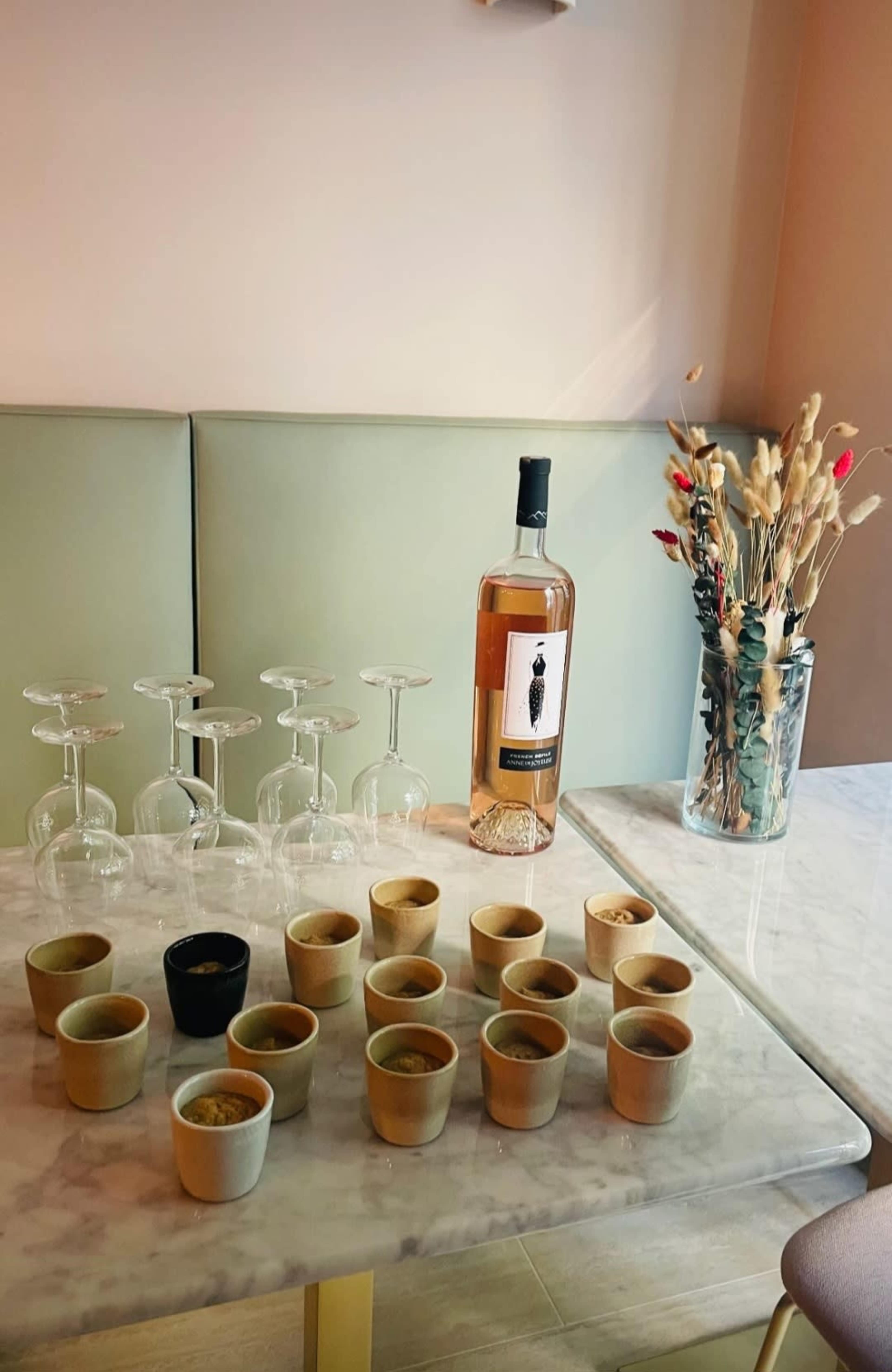 A bottle of rosé wine stands beside an arrangement of empty wine glasses and small ceramic cups on a marble table, with a vase of dried flowers in the background.