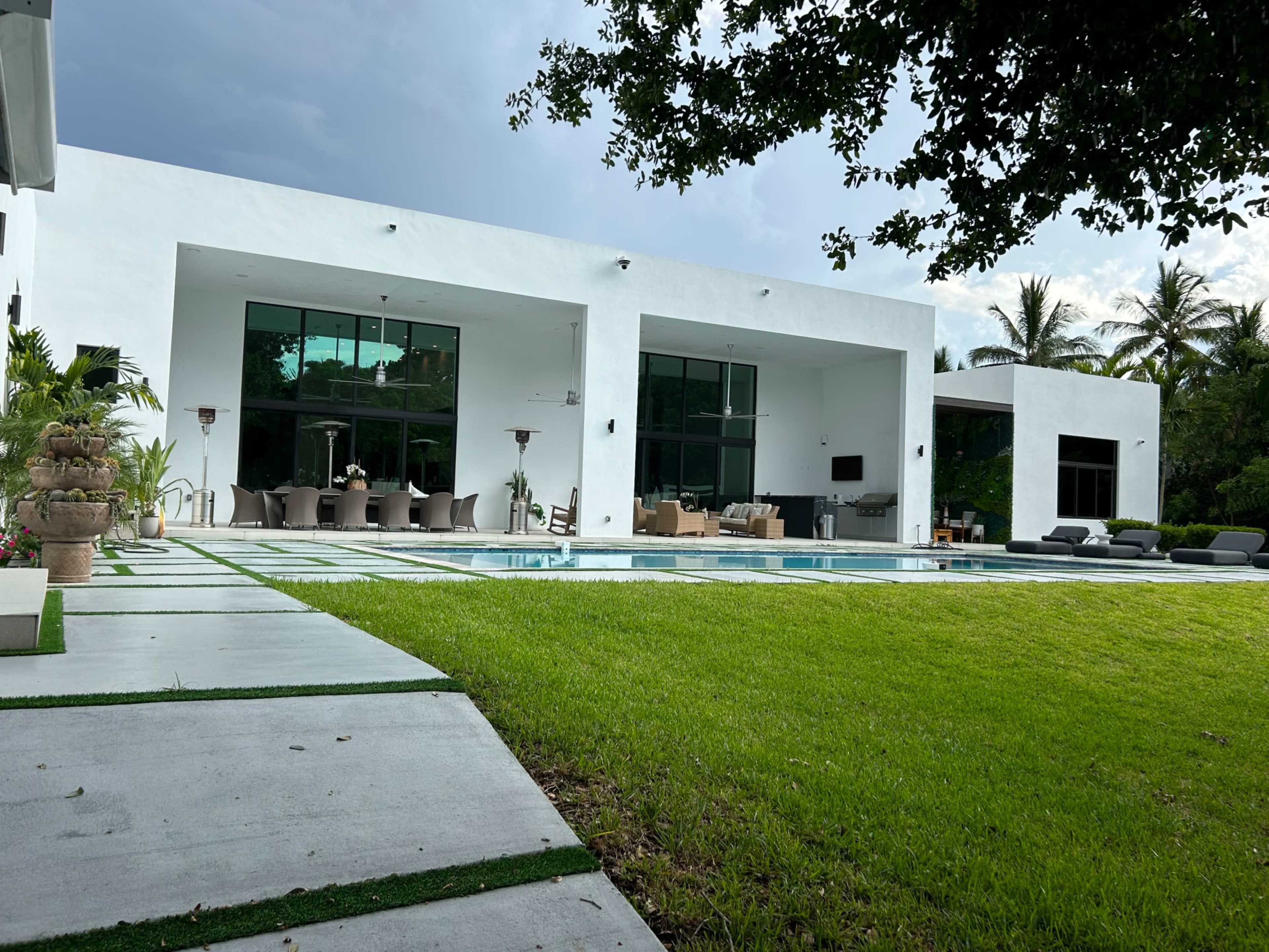 A modern white house with large windows and a swimming pool surrounded by a grassy area and palm trees.