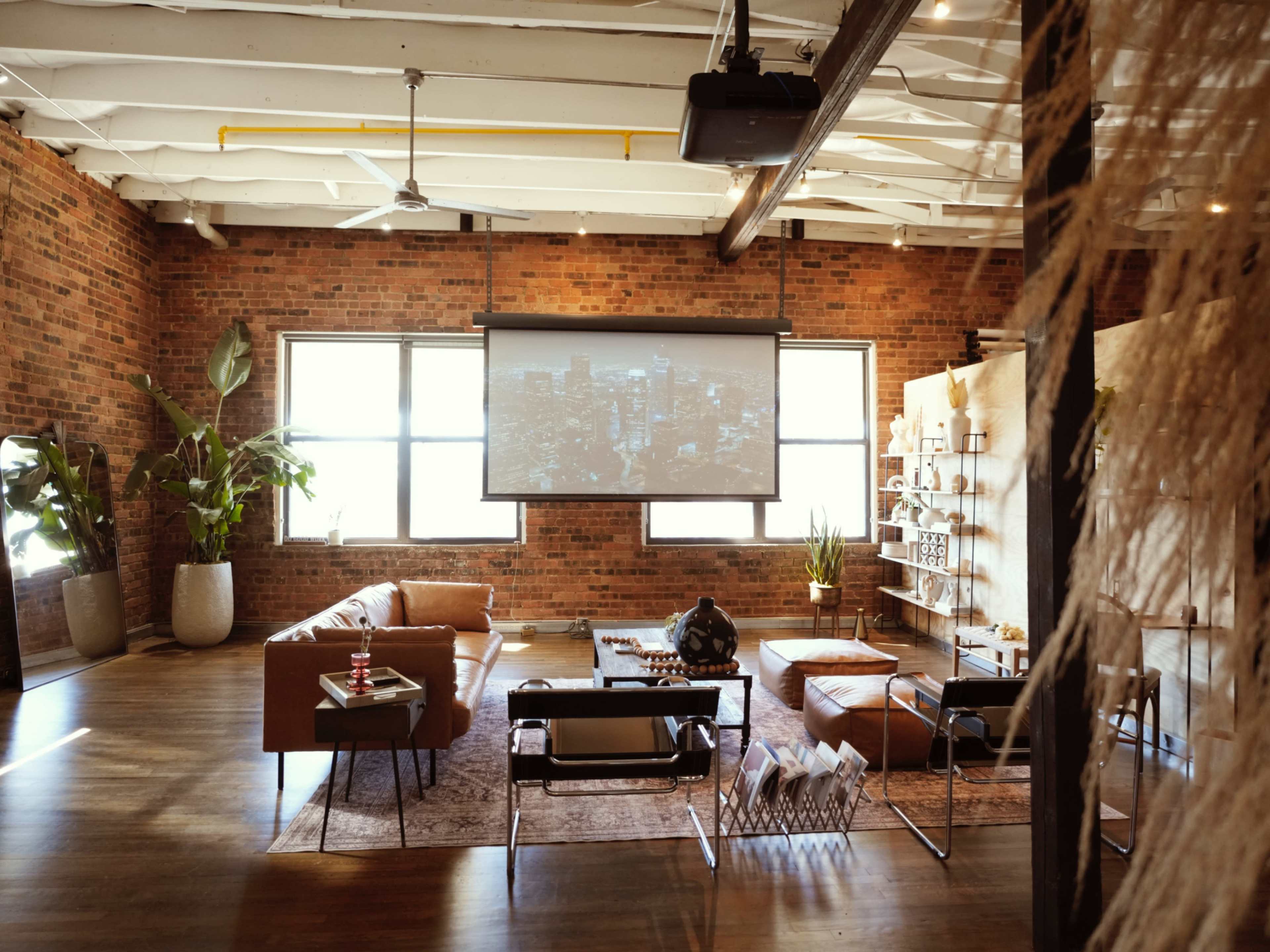 The image shows a modern loft-style living area with exposed brick walls, comfortable seating, a large projector screen, and decorative plants.